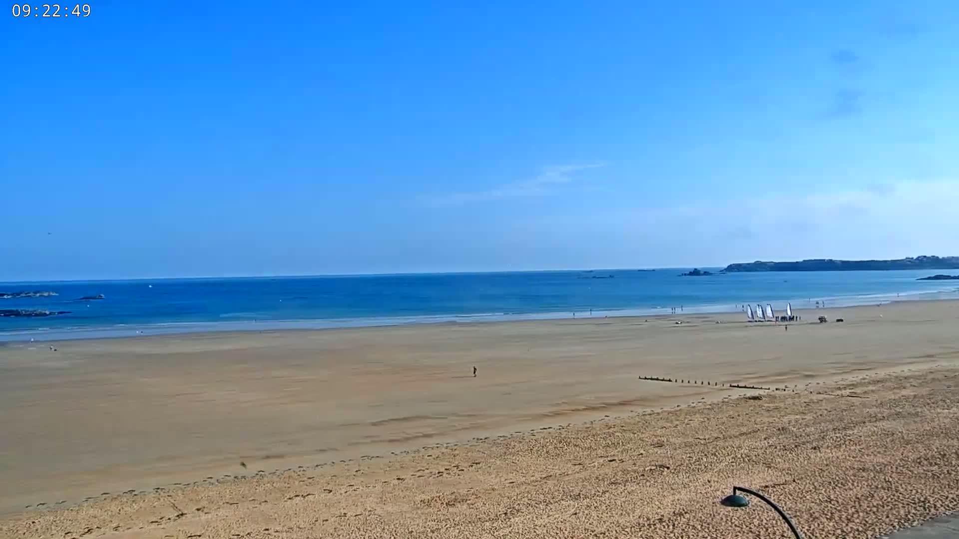 A wide sandy beach stretches to a calm, blue ocean under a clear blue sky, with a few people and sailboats visible in the distance.