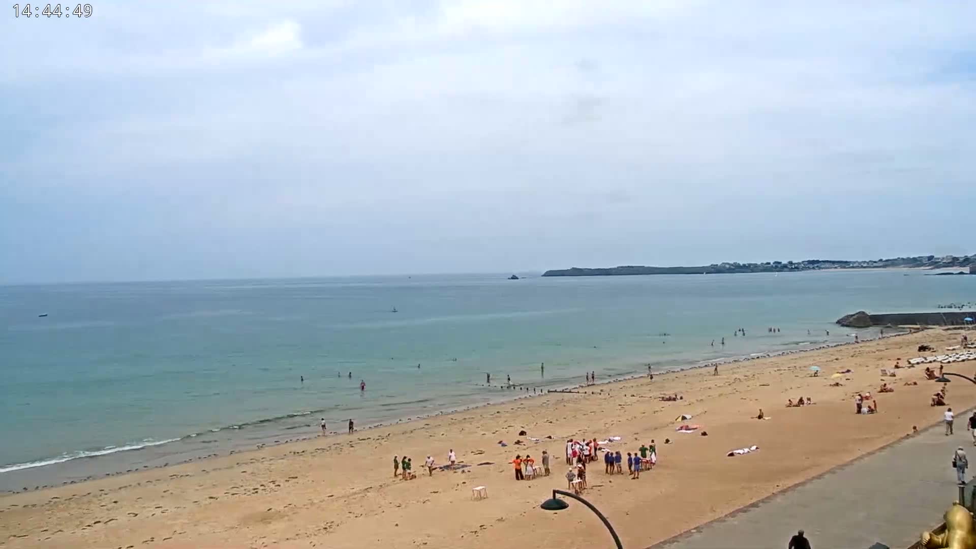 A sandy beach with scattered people is shown, next to a calm, light-blue ocean under a mostly cloudy sky.