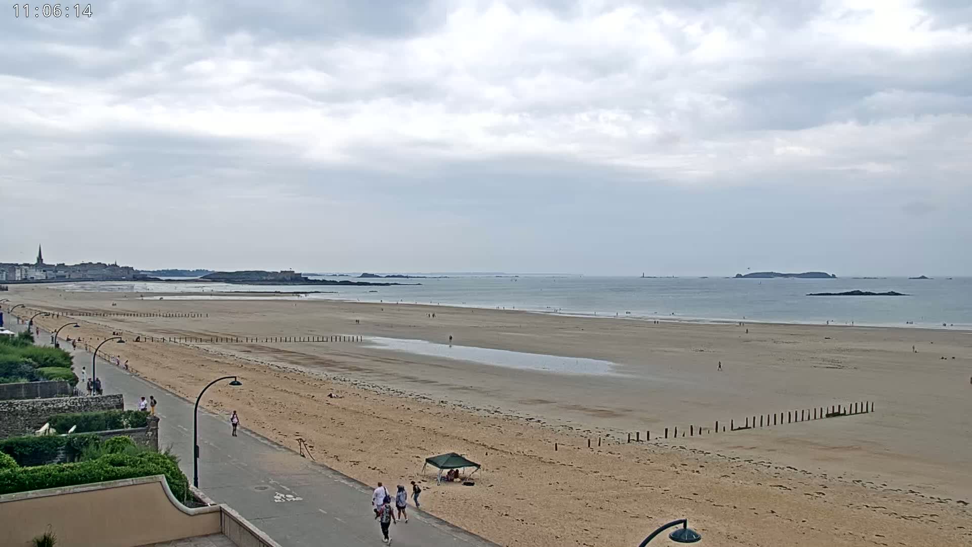 A wide sandy beach stretches to a calm sea under a cloudy sky, with a few people strolling along the shore and a town visible in the distance.