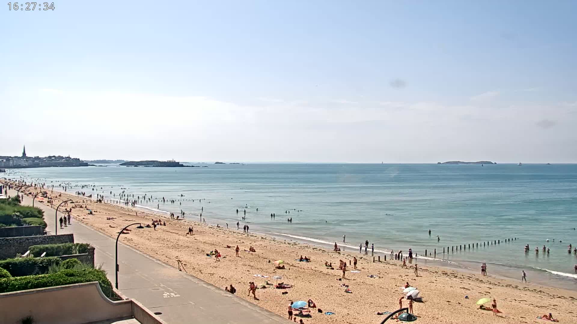 A sandy beach with many people swimming and sunbathing stretches along a calm, light-blue ocean under a clear, sunny sky.