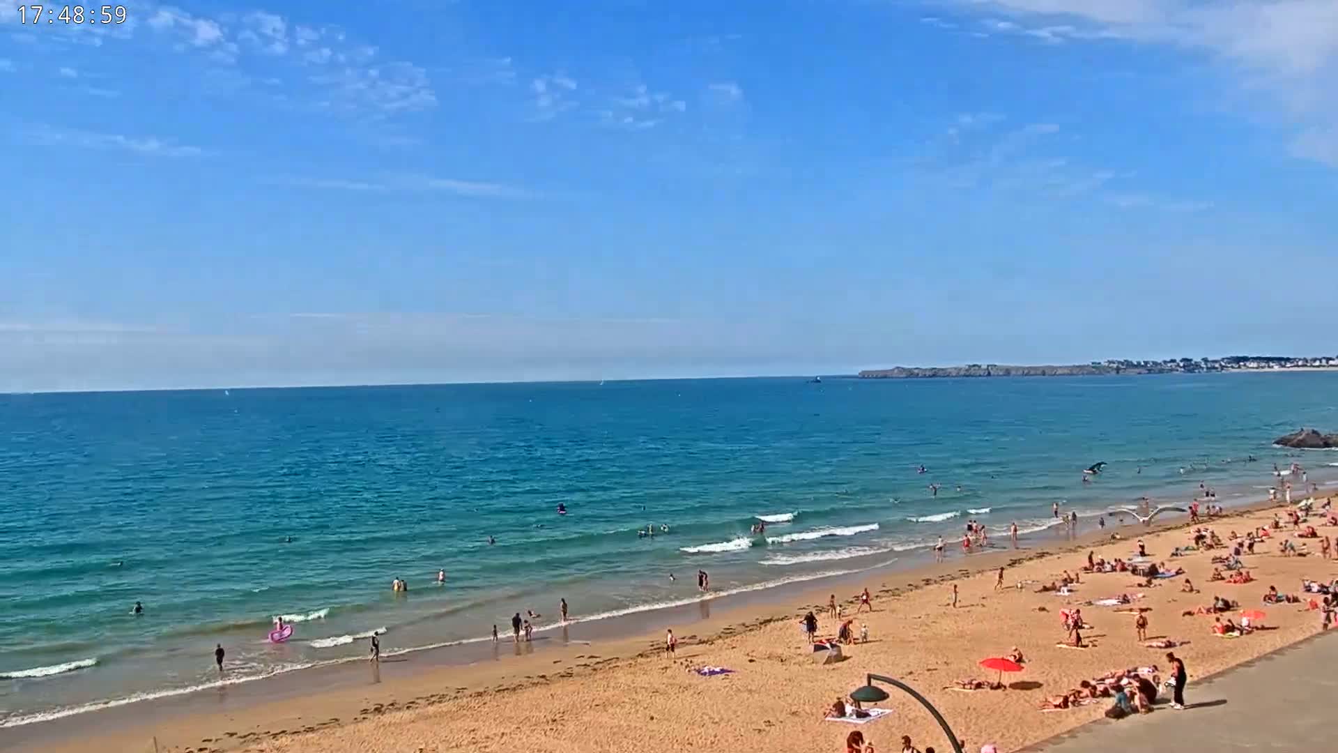 A sandy beach with many people sunbathing and swimming in a calm, blue ocean under a mostly clear, sunny sky.