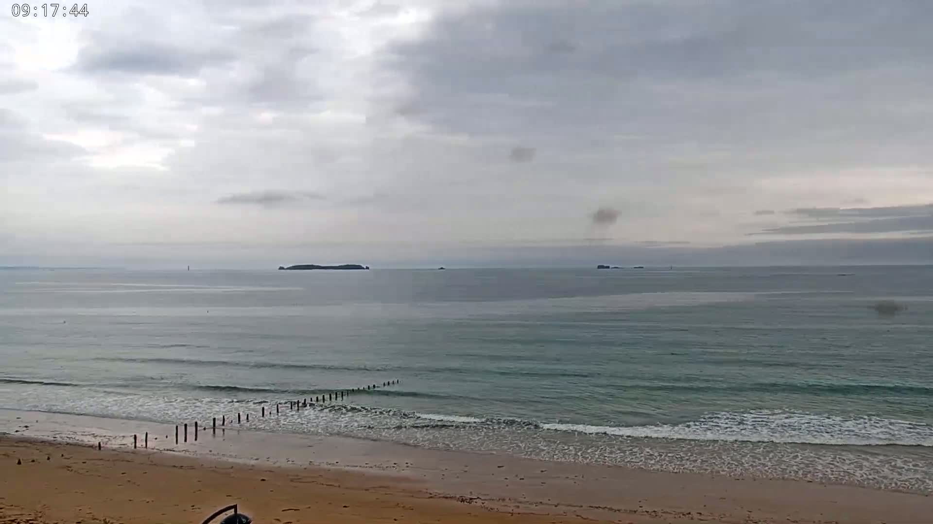 A sandy beach with small waves rolling onto the shore under a cloudy sky, with islands visible in the distance.