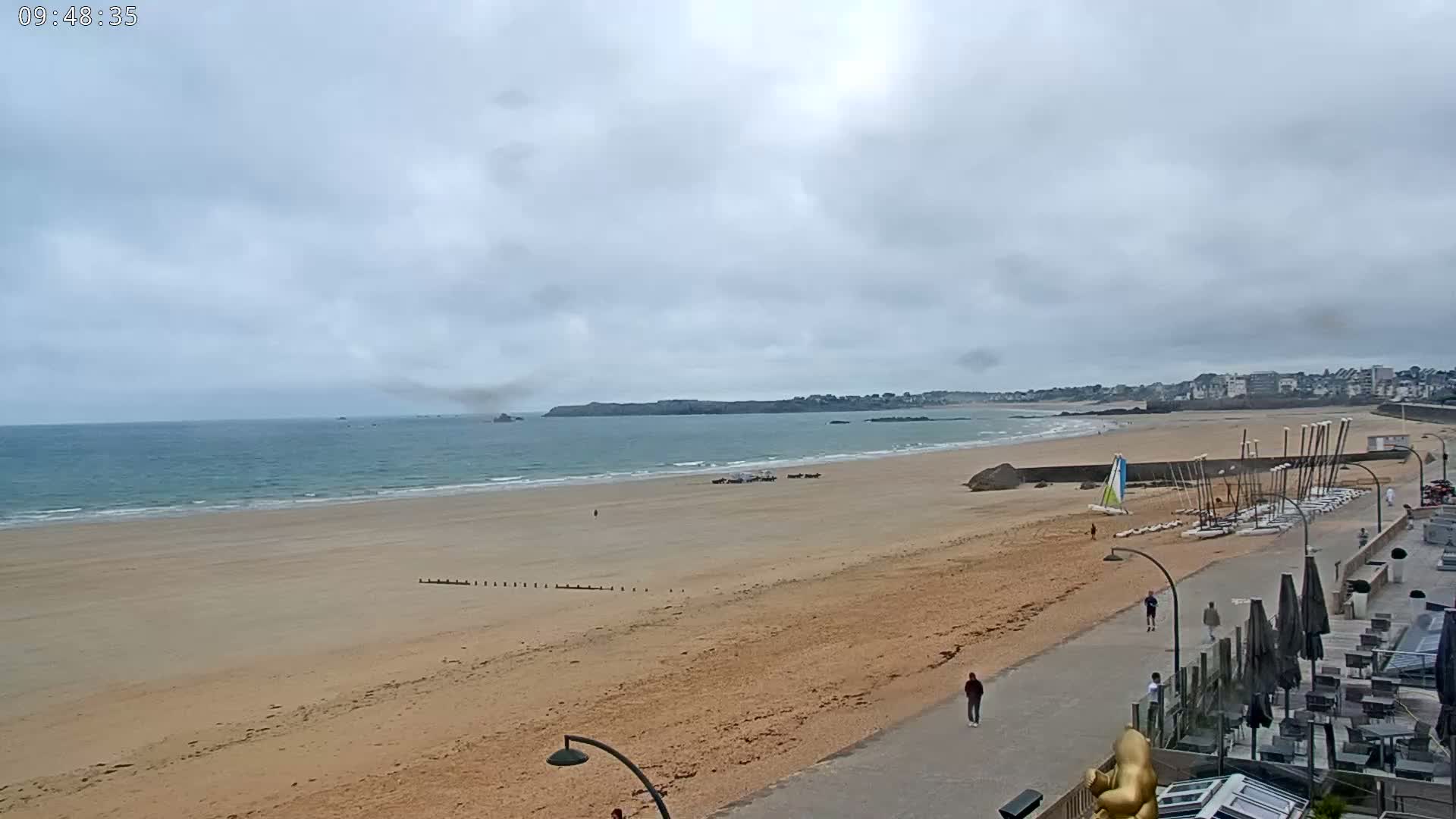 A wide sandy beach stretches along a calm ocean under an overcast sky, with sailboats stored on the shore near a paved walkway and buildings.
