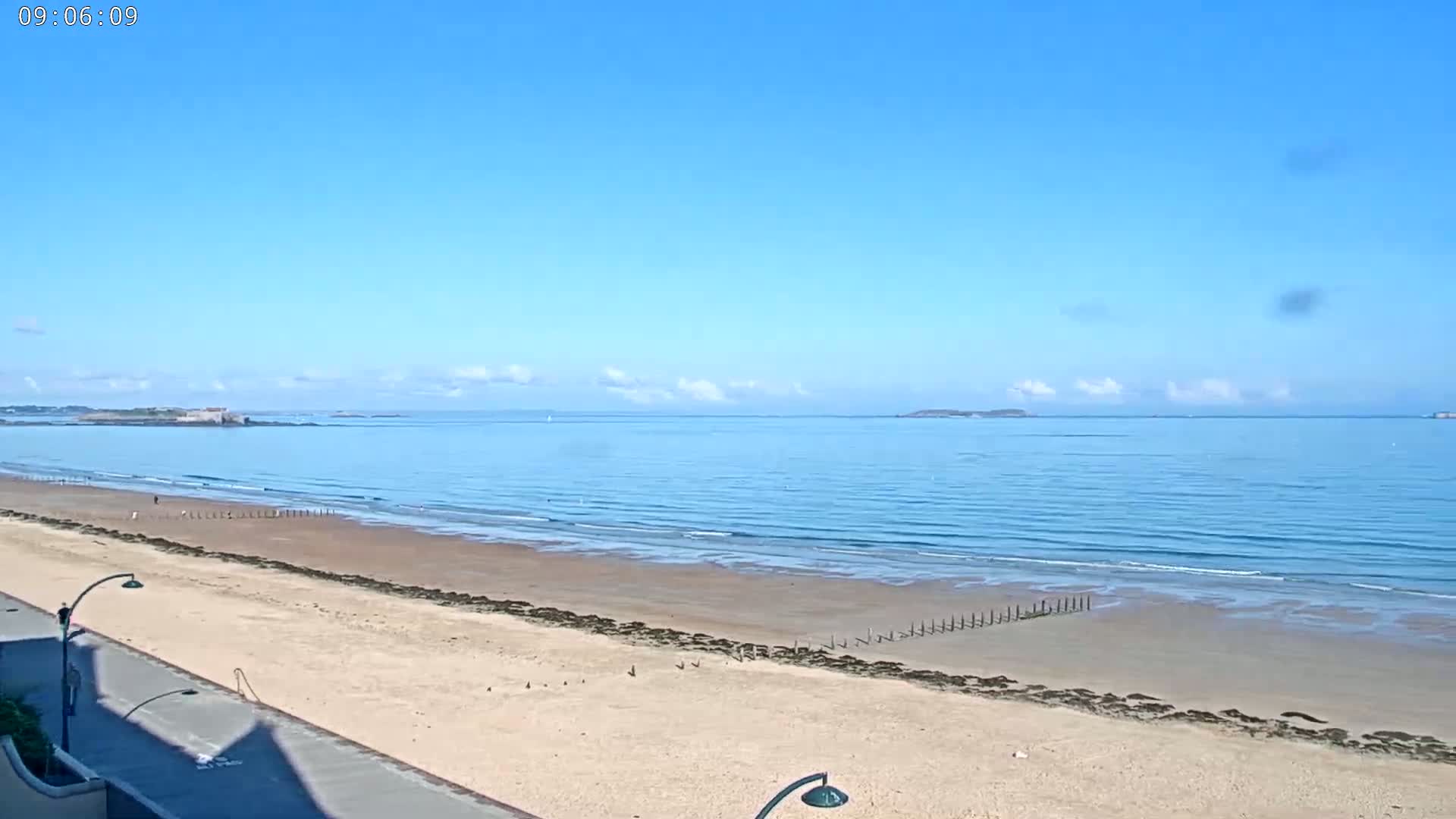 A sandy beach with calm, light-blue ocean stretches to the horizon under a clear blue sky with a few small clouds.