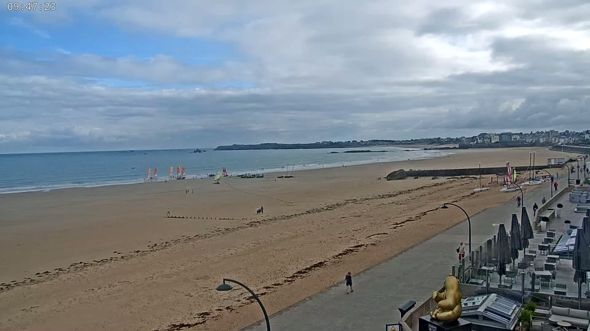 A wide sandy beach with several sailboats on the water under a partly cloudy sky, and a paved walkway with outdoor seating along the beach.