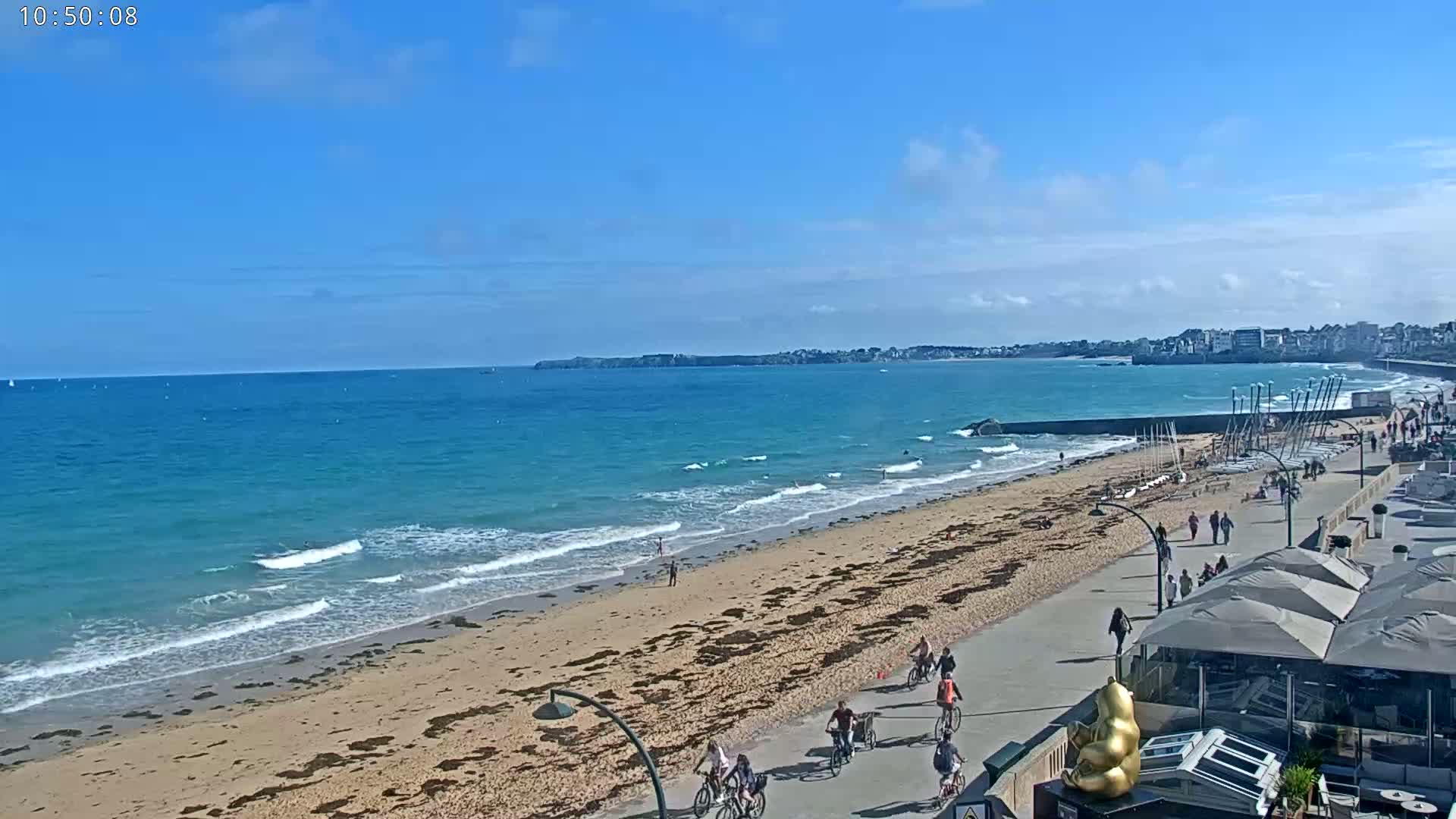 A sandy beach with gentle waves under a mostly clear blue sky, showing people walking, biking, and enjoying the outdoors near a seaside town.