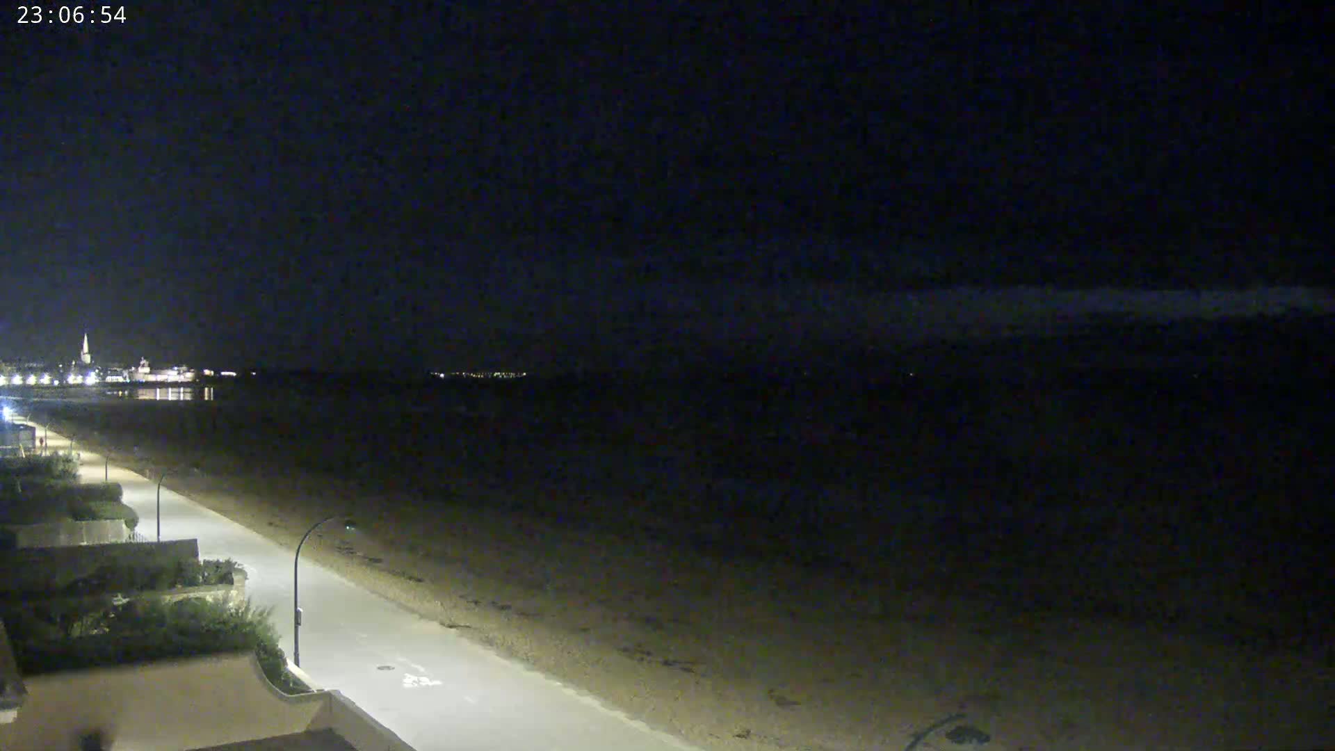 A nighttime view of a mostly empty beach and road leading to a dimly lit town across the water under a dark sky.
