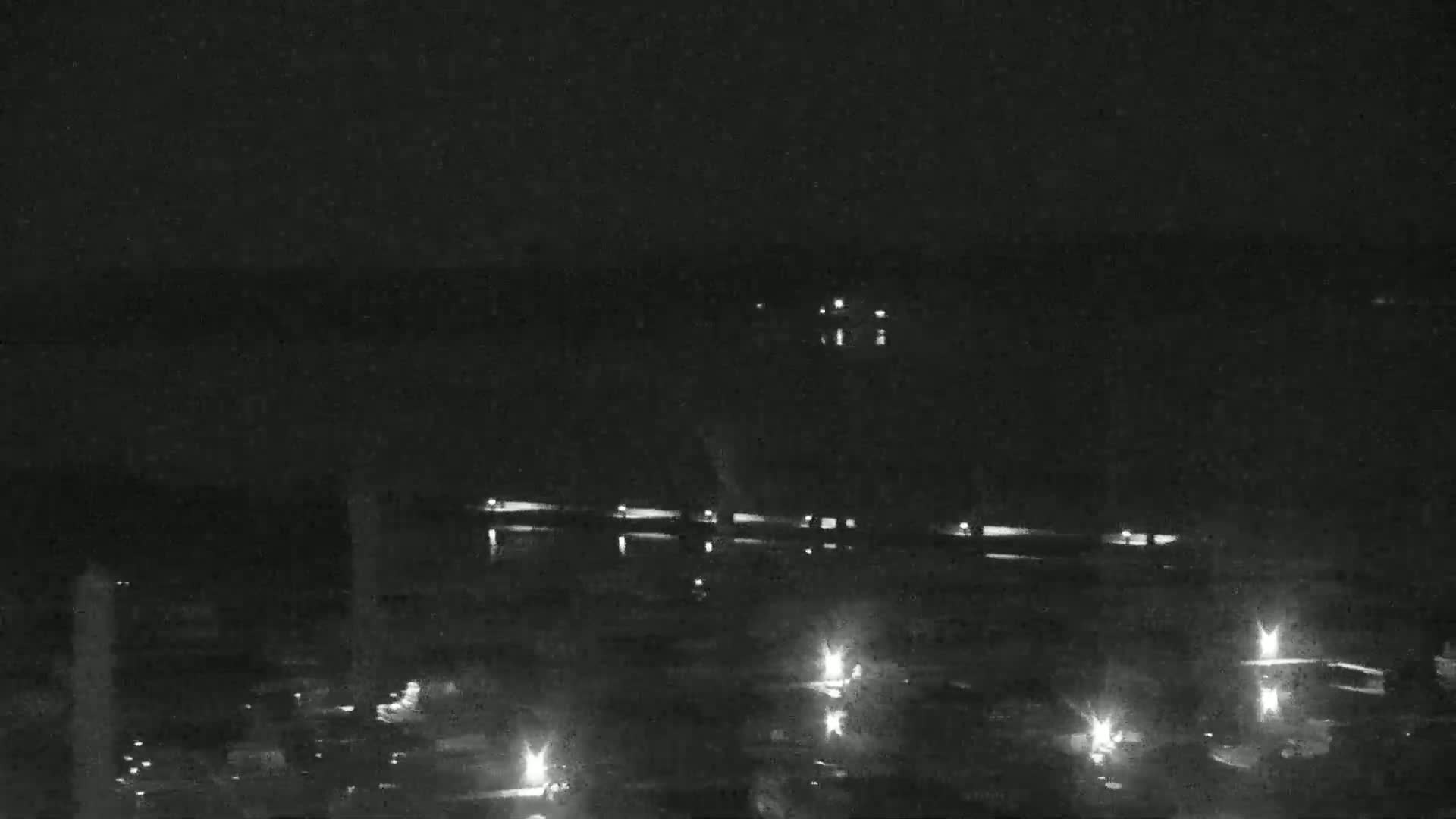 A nighttime, low-light view shows a long, dark structure with scattered lights, possibly a building or bridge, situated near a body of water under a clear, dark sky.