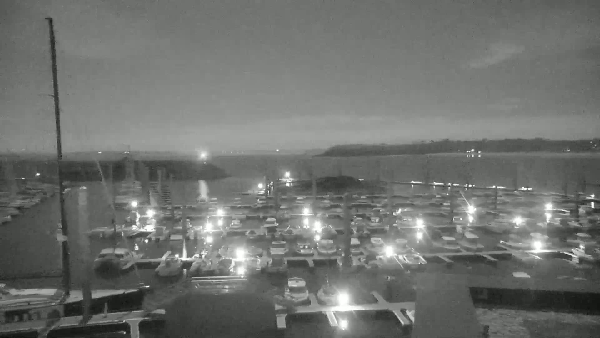 A nighttime, low-light view of a marina filled with numerous boats illuminated by small lights, situated on a body of water under a mostly clear sky.