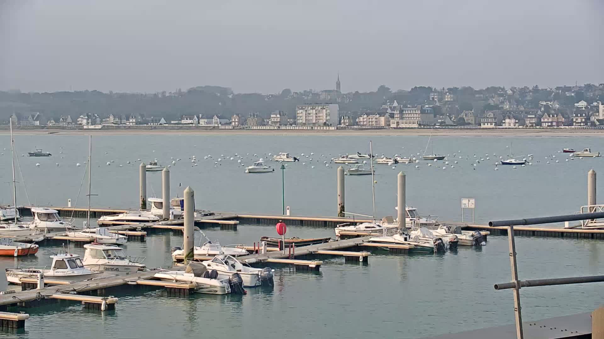 Many boats are docked at a marina, with additional boats and buoys in the calm water of a bay, and a town visible in the distance under a hazy sky.
