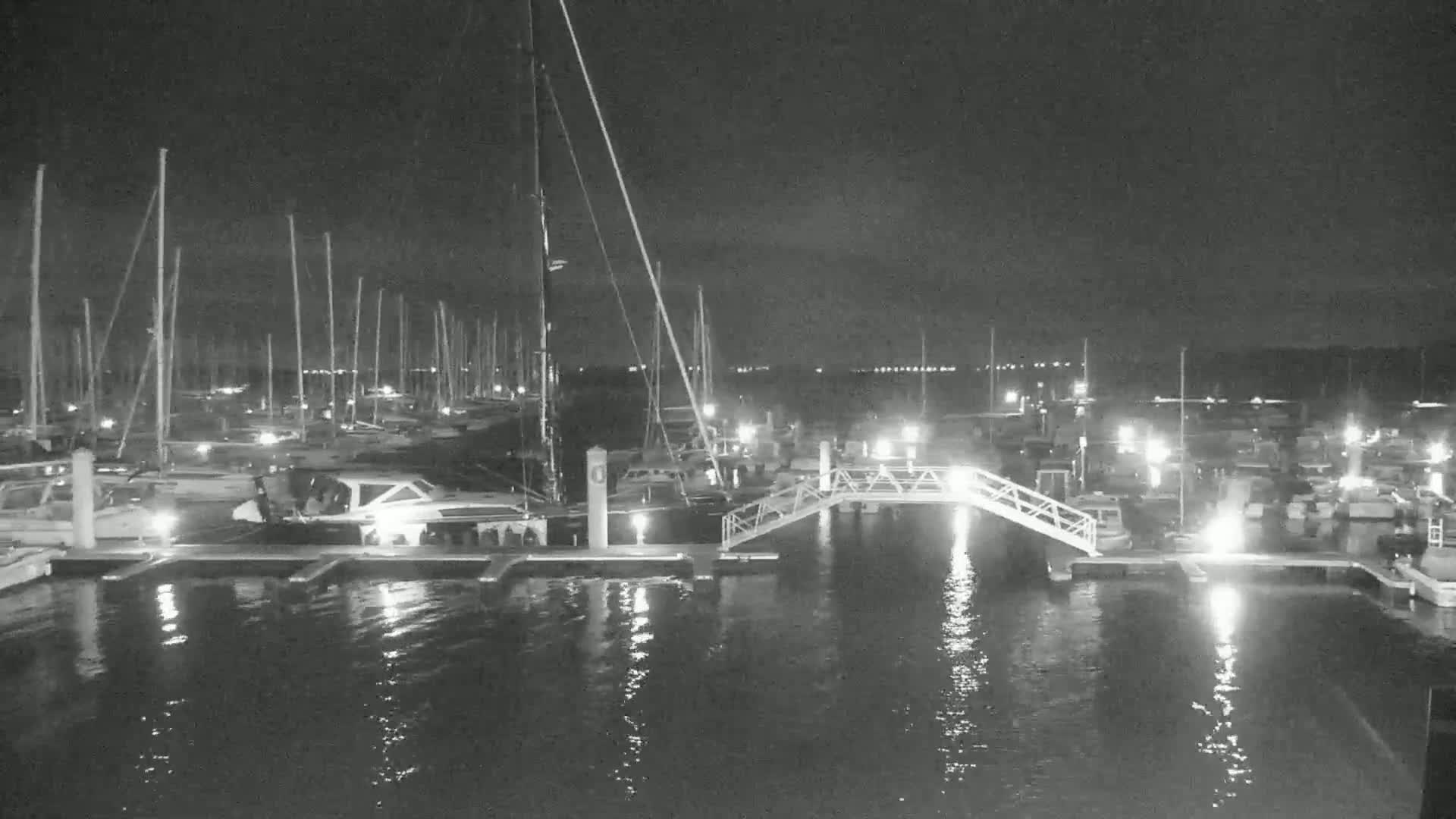 A nighttime view of a marina with many boats docked, illuminated by lights, and a calm, reflective body of water.