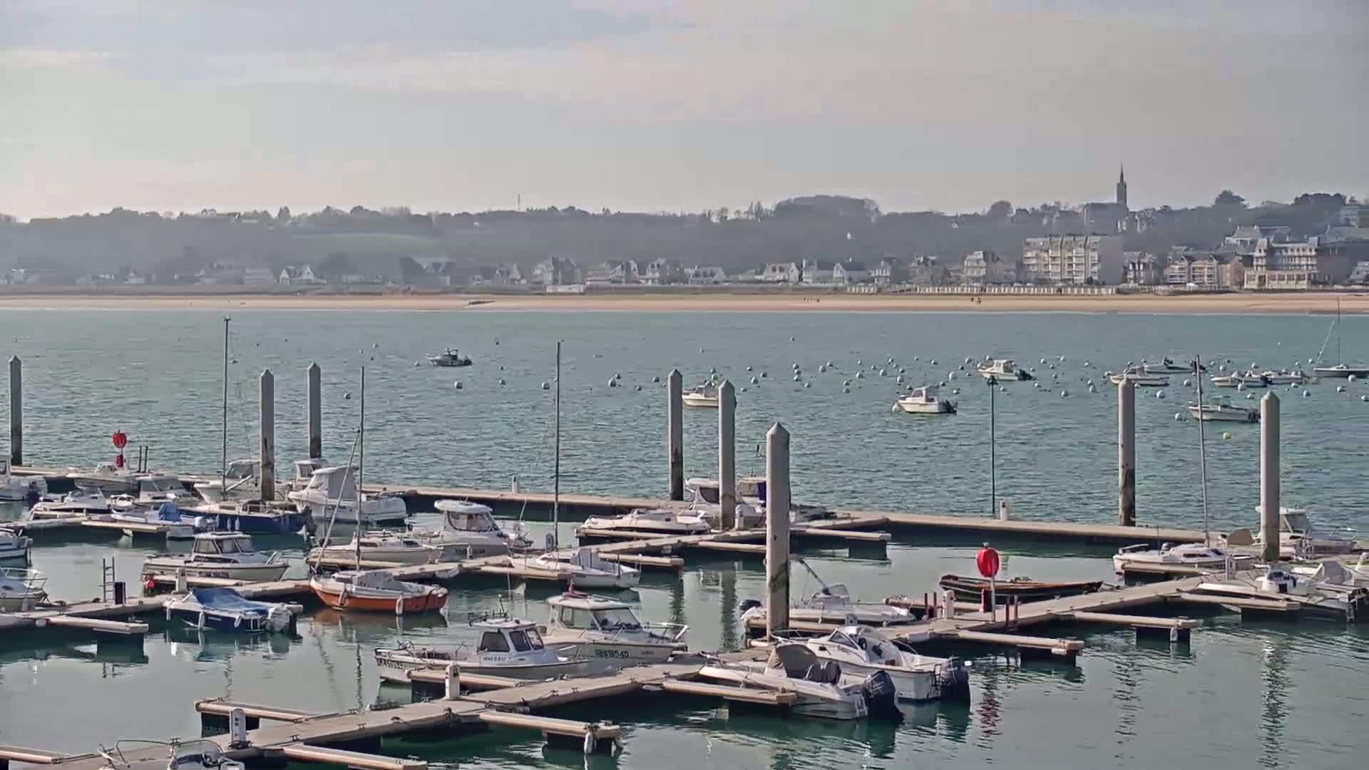Numerous boats are moored at a marina, with a town and sandy beach visible across calm water under a bright, clear sky.