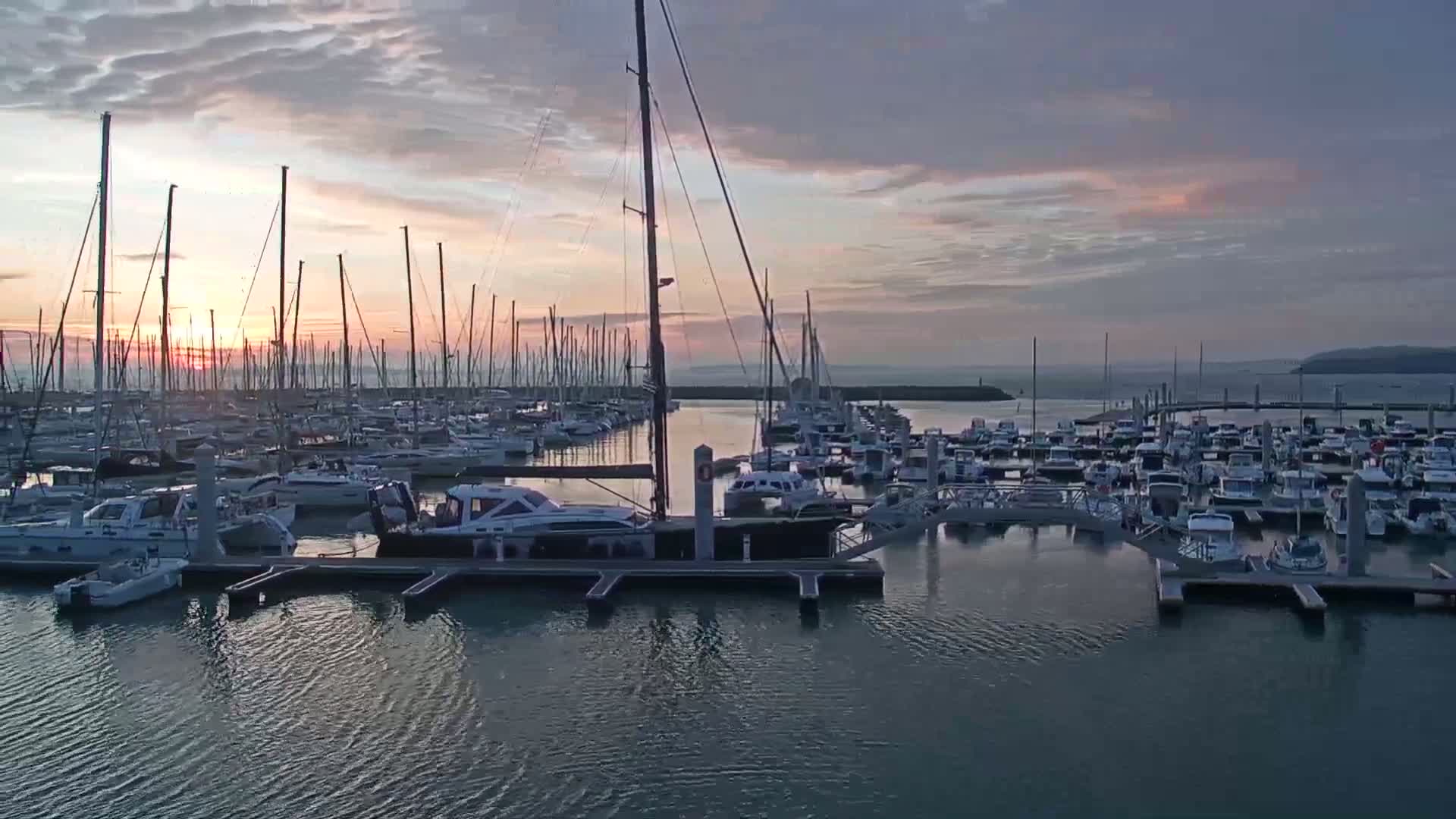 Many sailboats and motorboats are docked in a marina at sunrise under a partly cloudy sky.