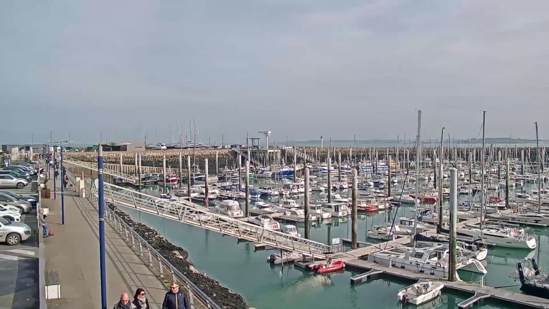 A marina filled with numerous boats and yachts is seen on a sunny day, with people walking along a paved walkway next to a parking lot.