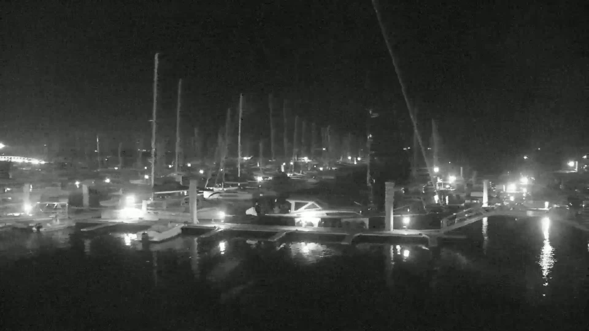 A nighttime view of a marina with numerous boats docked, illuminated by lights, reflecting on calm water.