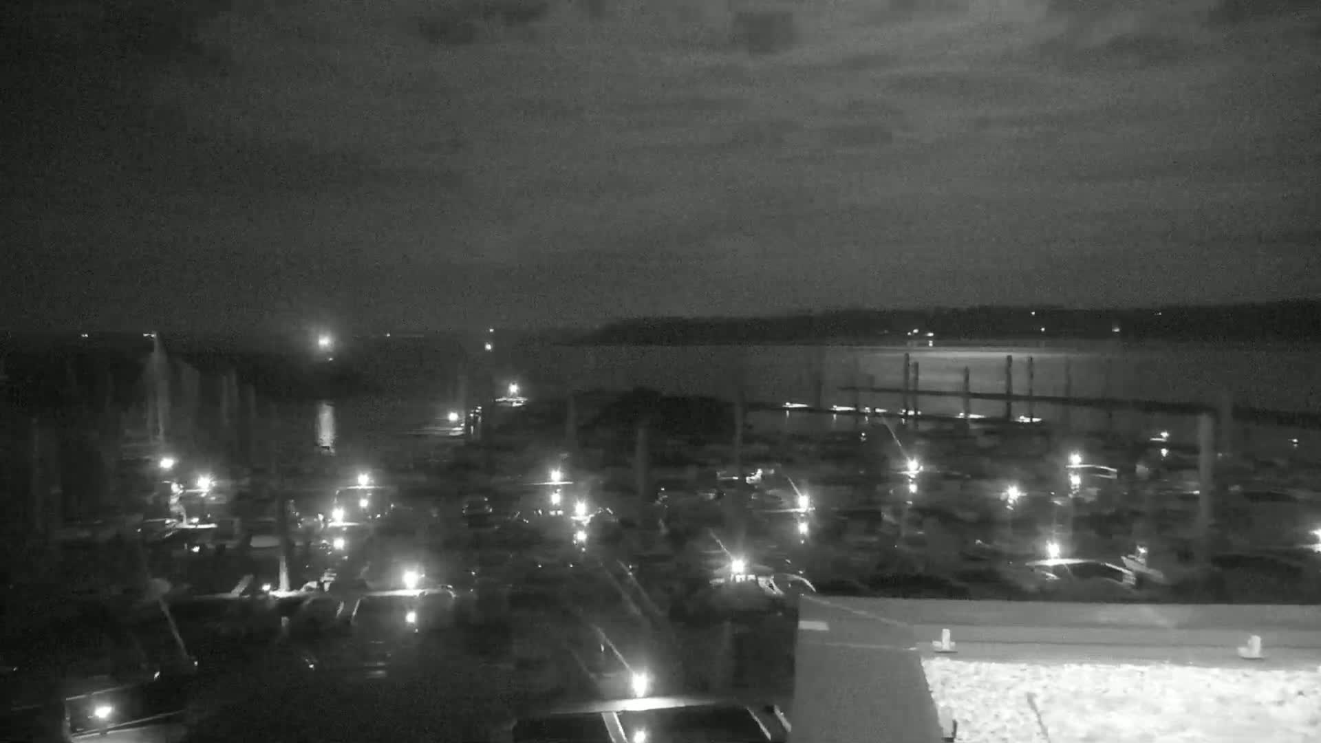 A nighttime view of a marina filled with boats, illuminated by their lights, overlooking a calm body of water under a dark sky.
