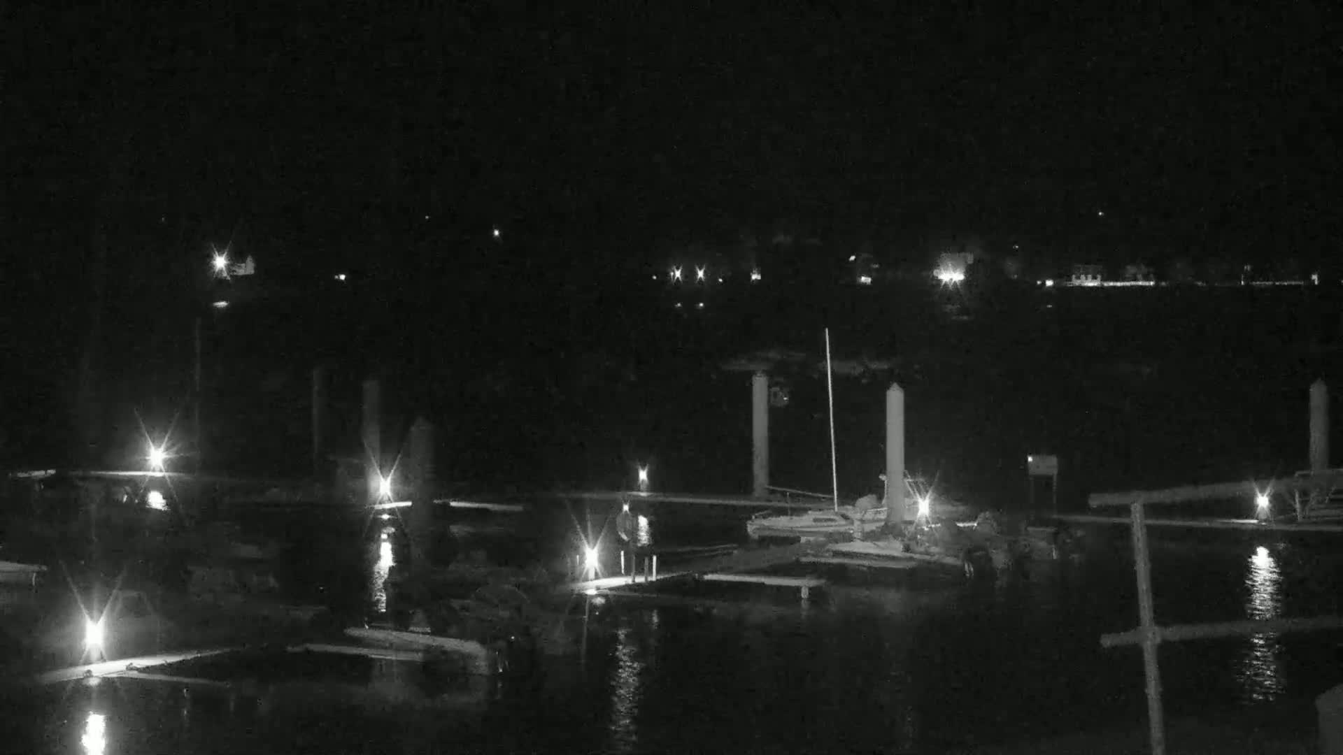 A nighttime view of a marina with boats docked, illuminated by various lights, under a clear, dark sky.