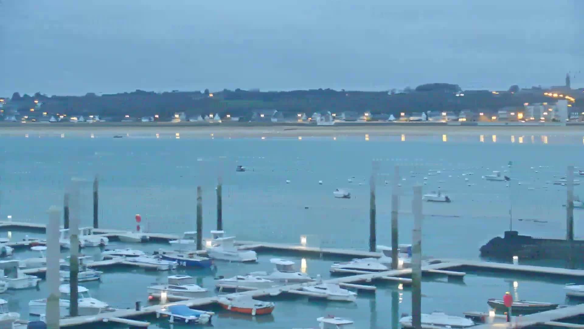 A marina filled with numerous boats is situated in calm, twilight waters before a distant town illuminated at night.