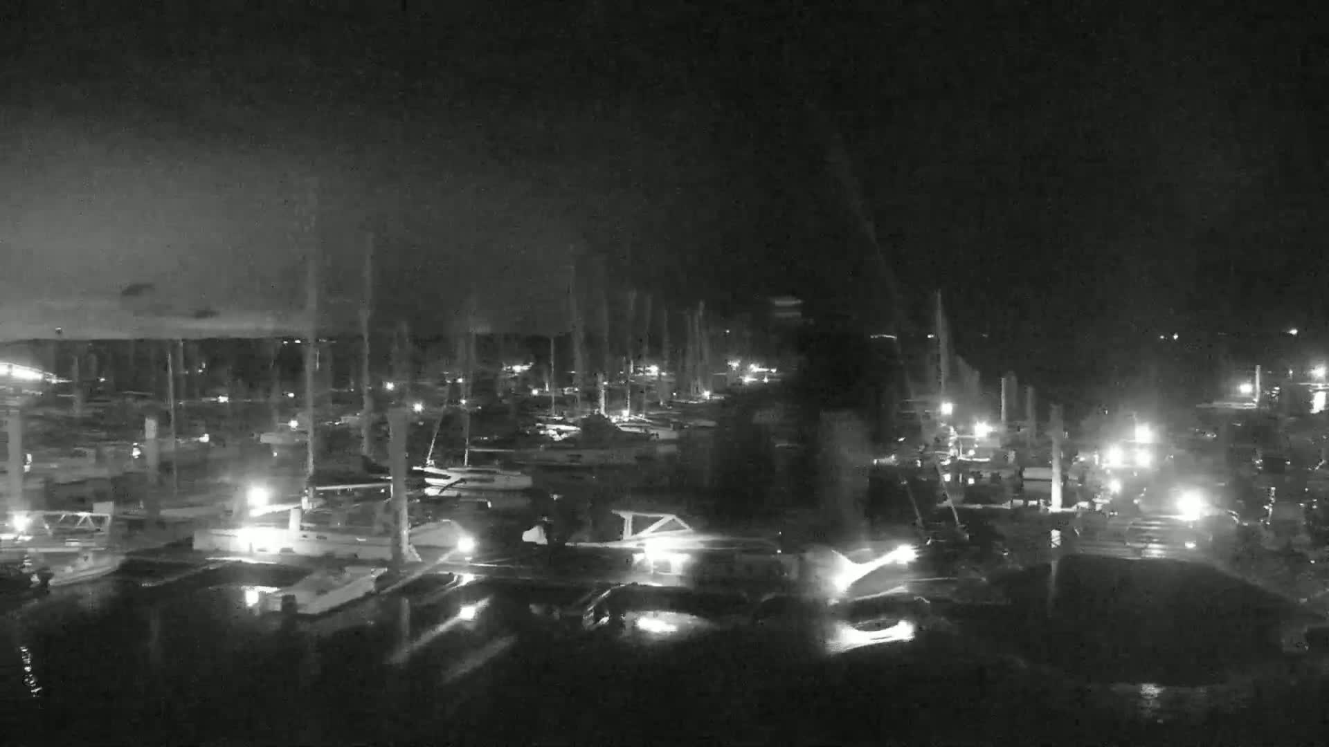 A nighttime, low-light view of a marina filled with numerous boats, their masts and lights visible under a dark, possibly cloudy sky.