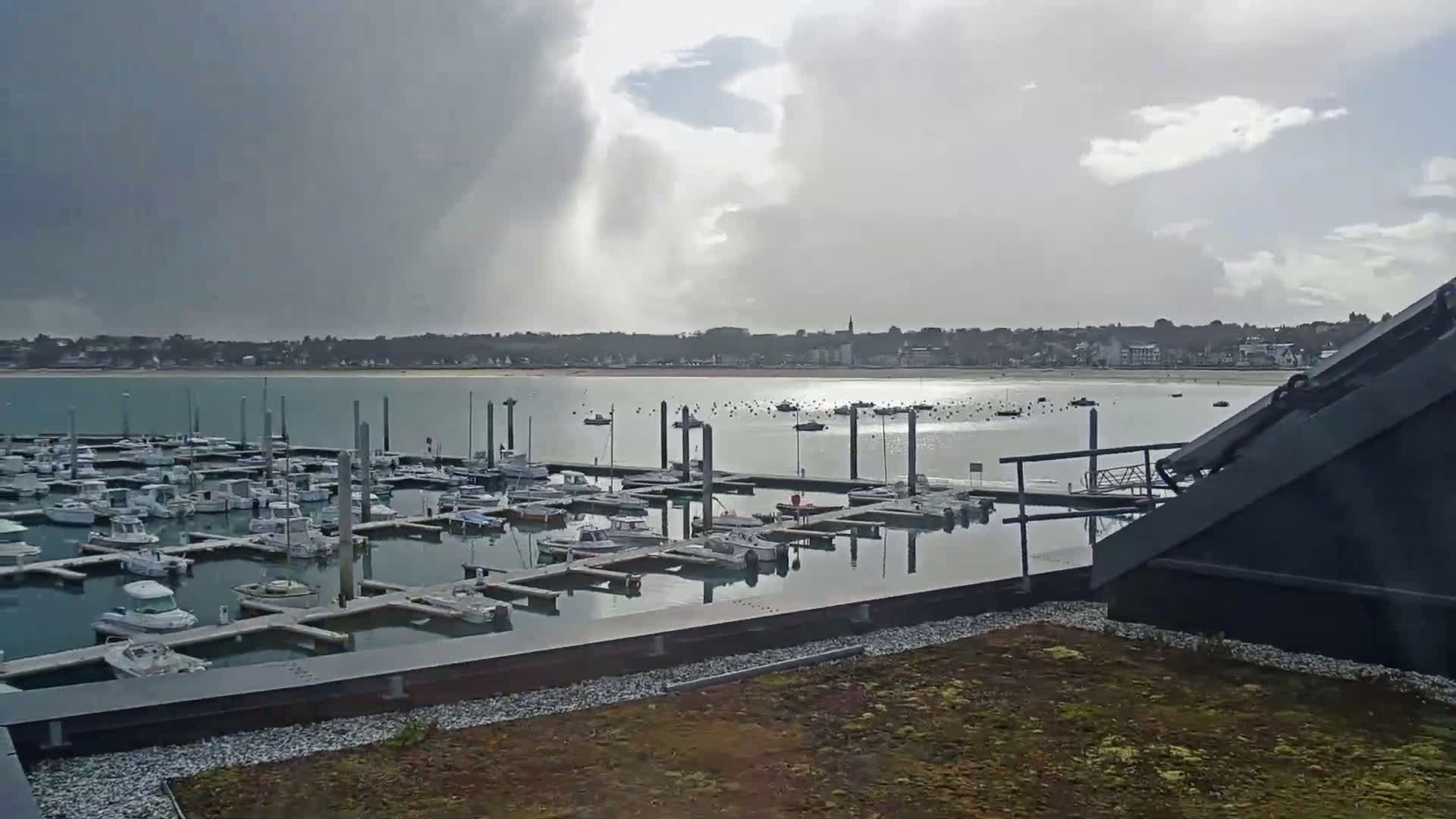 A marina filled with numerous boats is seen under a partly cloudy sky, with a town visible in the background across a body of water.
