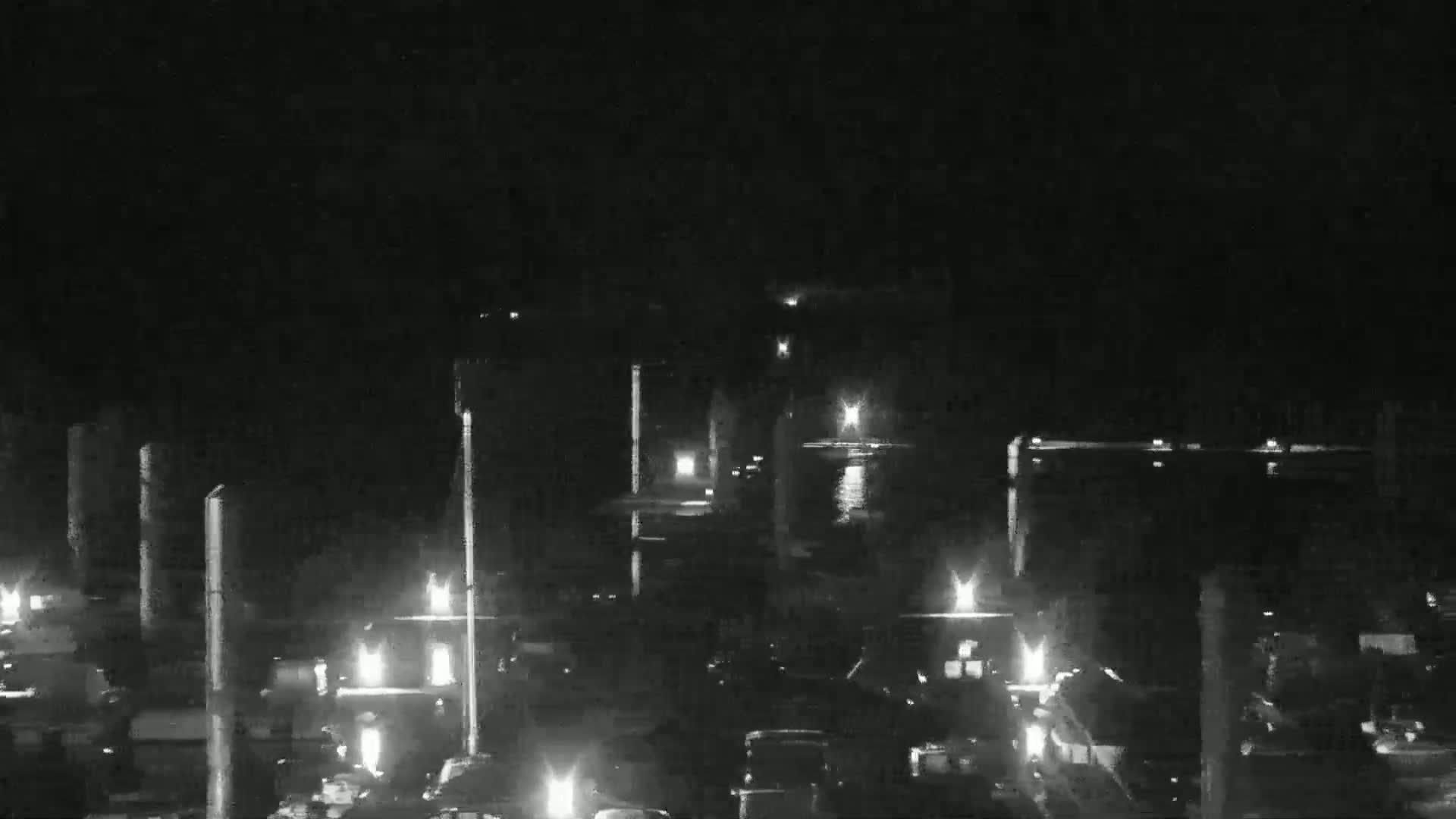 A nighttime, low-light view of a waterfront area with numerous lights illuminating vehicles and structures along a calm body of water.