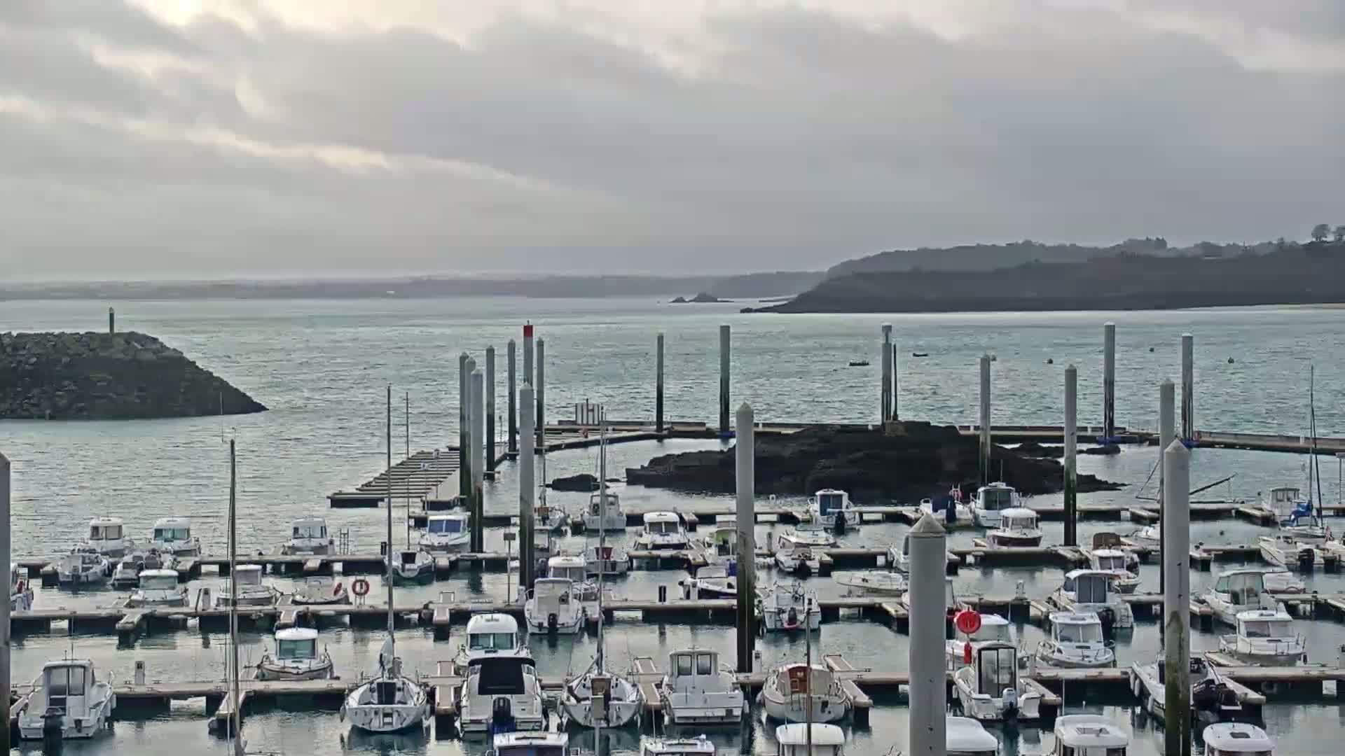 A marina filled with numerous boats is shown under a cloudy sky, with calm water visible beyond the harbor.
