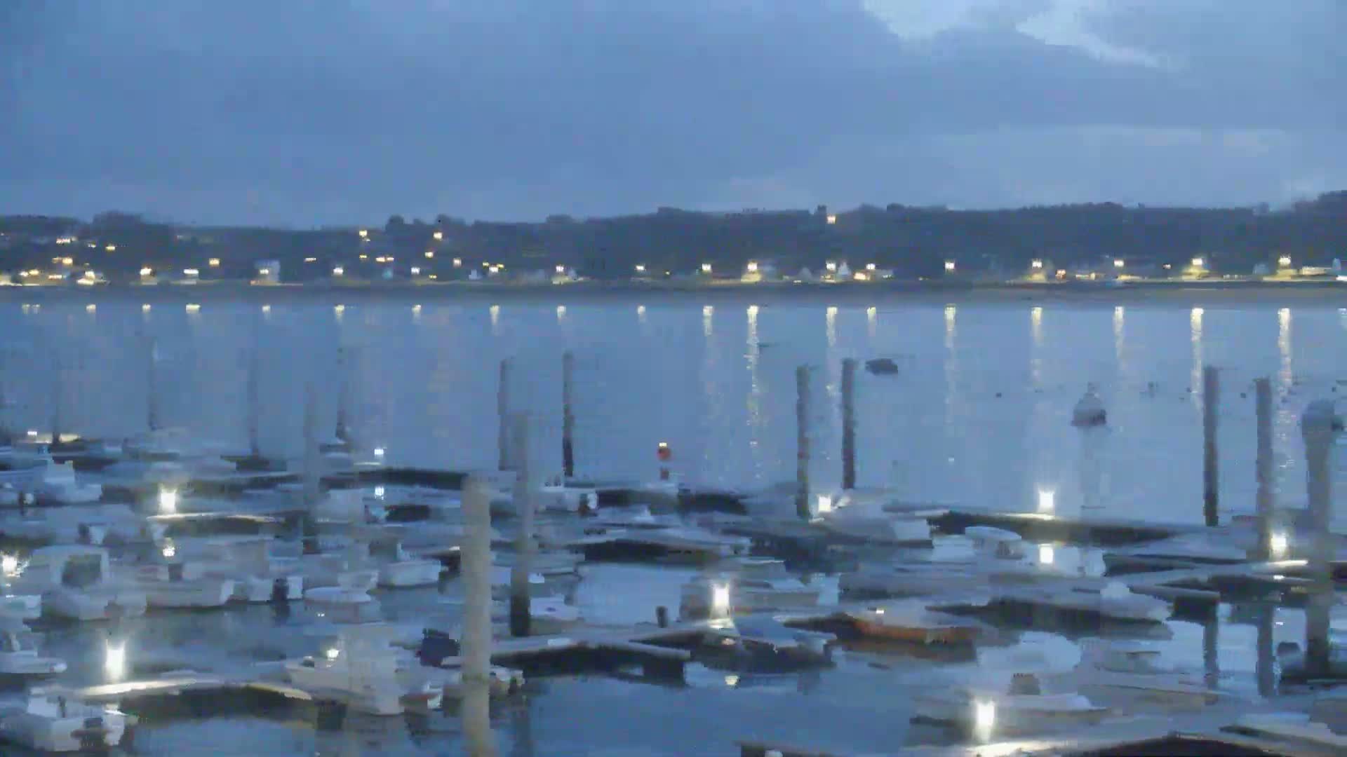 Many boats are docked at a marina at twilight, with a calm body of water reflecting the lights of a distant town.