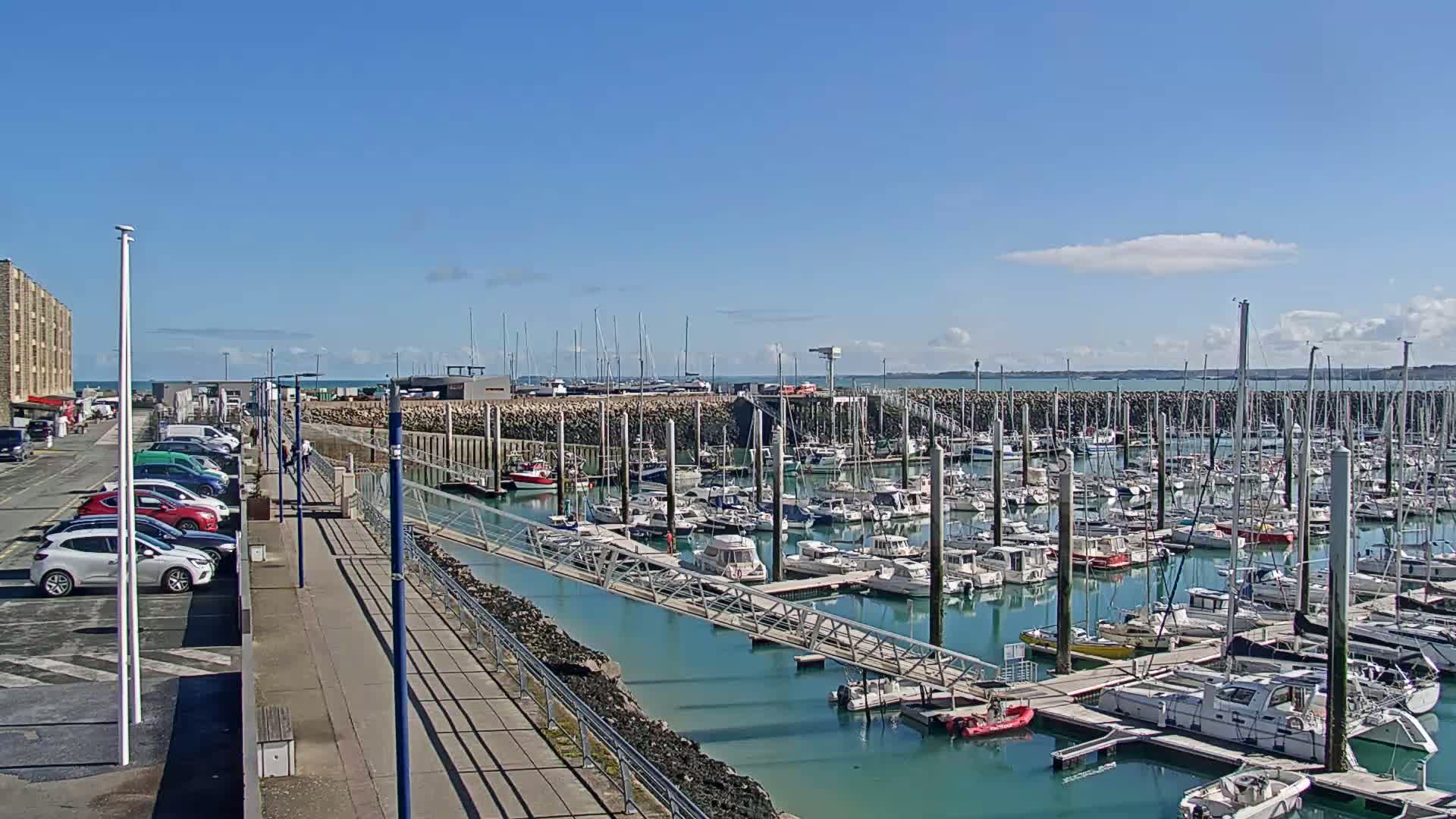 A marina filled with numerous boats and yachts is shown on a sunny day, with a parking lot and walkway visible nearby.