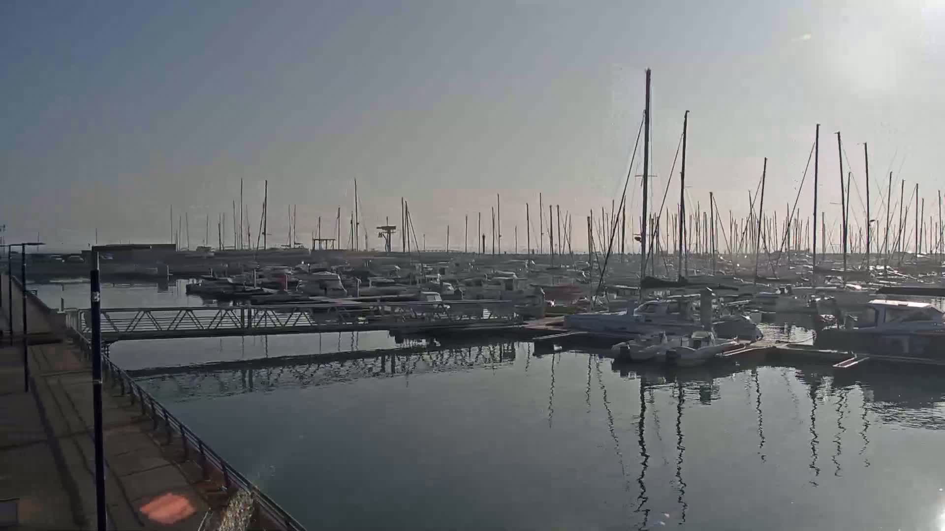 A calm marina filled with numerous sailboats and motorboats under a clear, light sky.