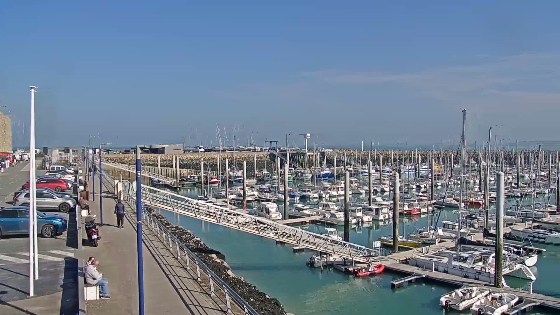 A sunny day at a marina filled with numerous sailboats and motorboats, with a walkway and parking lot alongside.