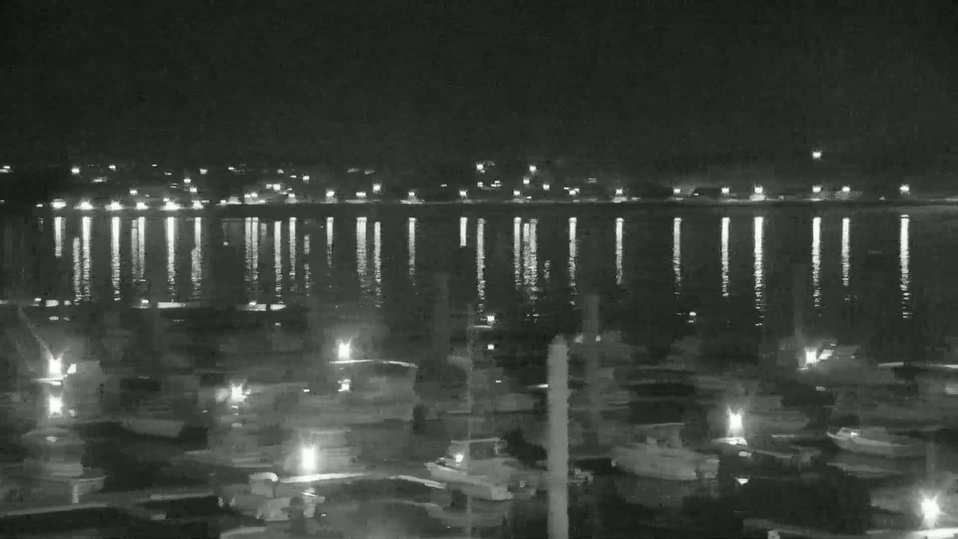 A nighttime view of a calm body of water reflecting lights from a town across the water, with numerous boats docked in the foreground.