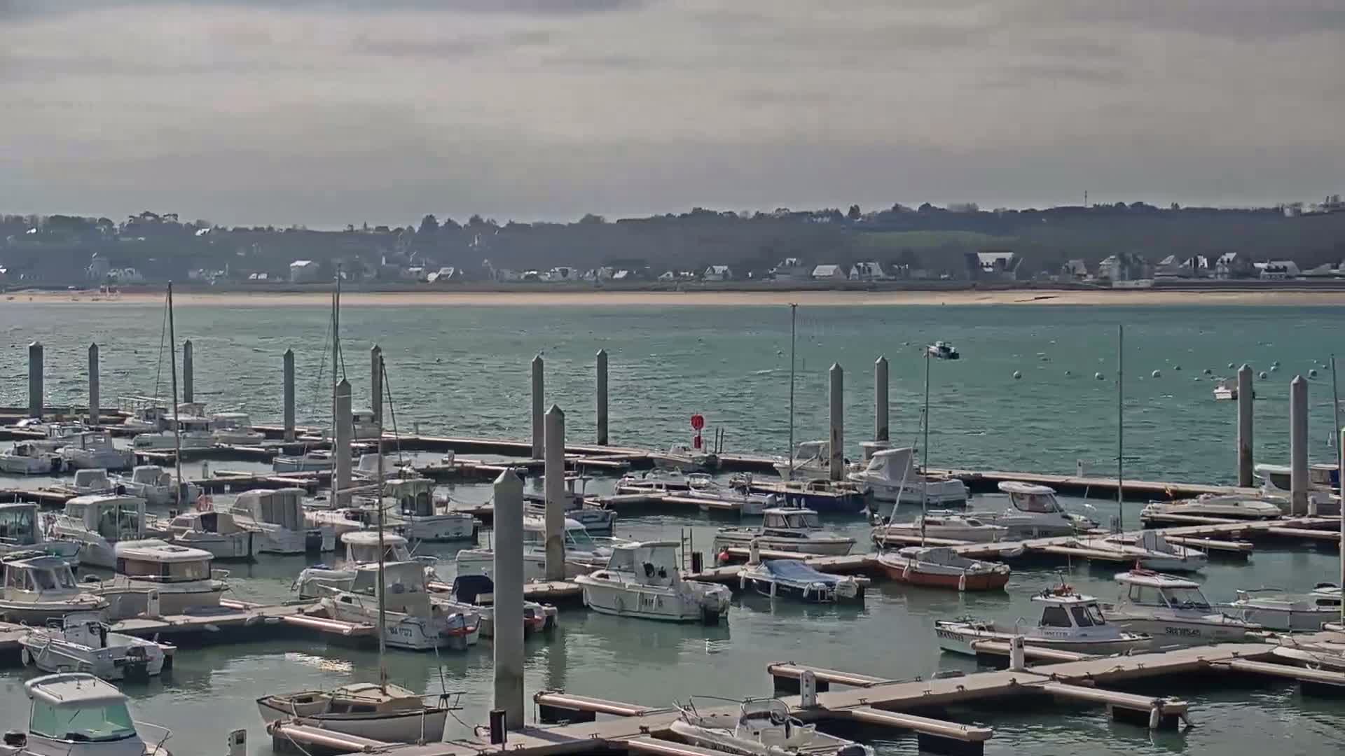 A marina filled with numerous boats is situated on a body of water under a cloudy sky, with a distant shoreline visible in the background.