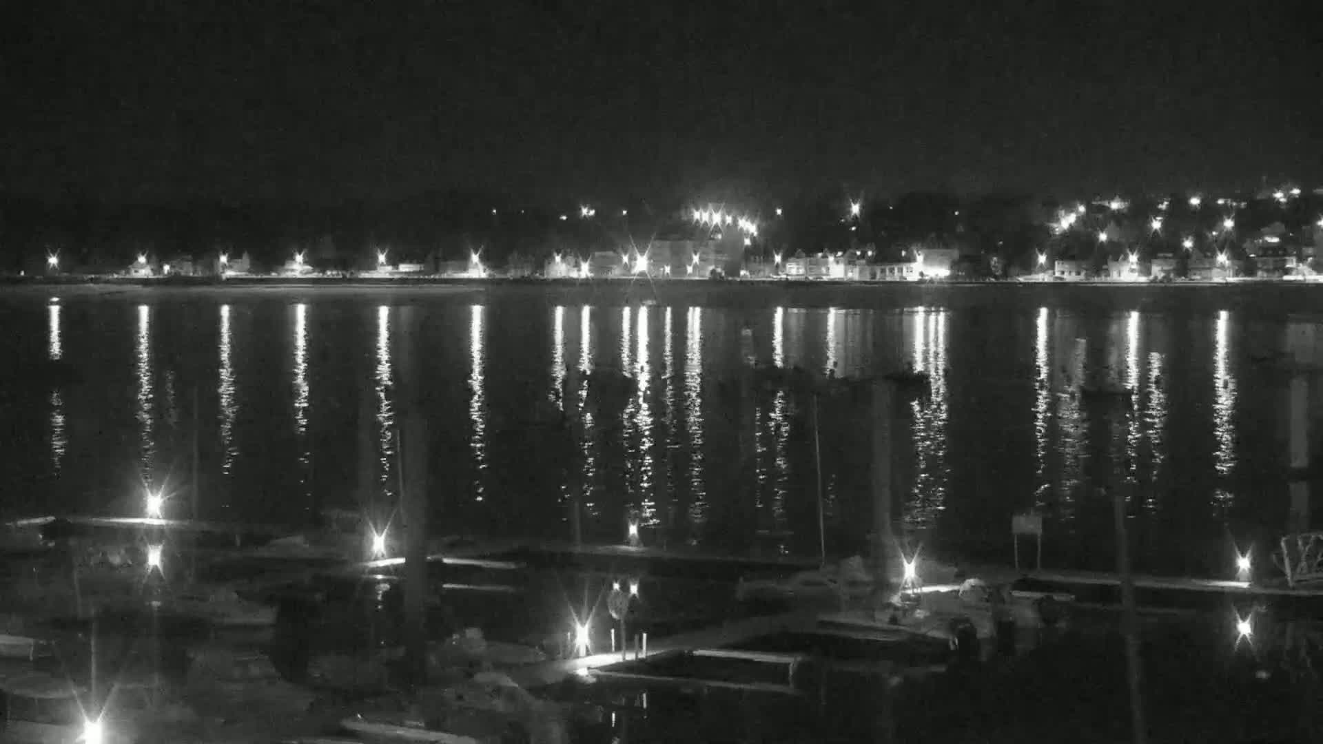 A nighttime view of a calm body of water reflecting the lights of a town across the shore, with numerous boats moored at a dock in the foreground.