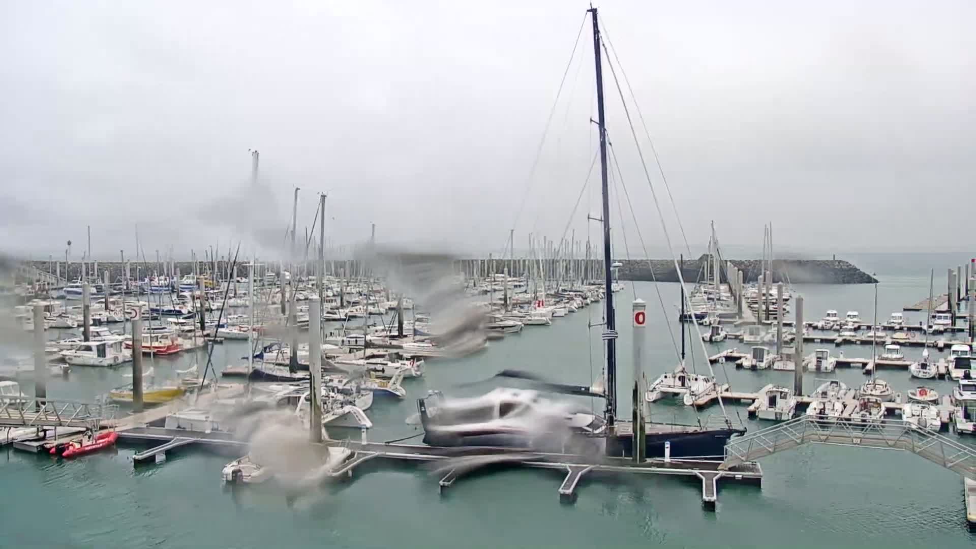 A marina filled with numerous sailboats and motorboats is seen in rainy weather.