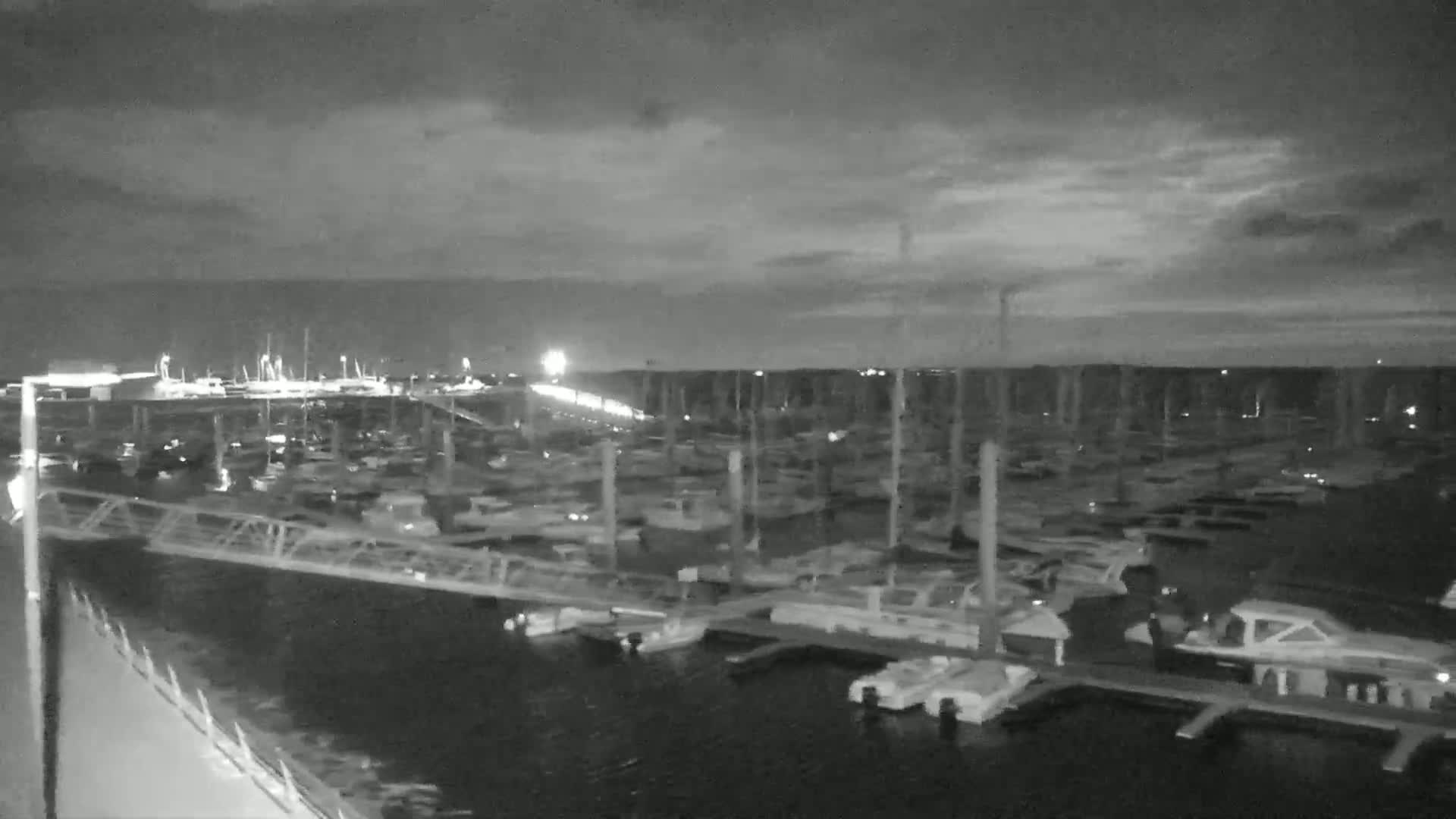 A nighttime, overcast view of a marina filled with numerous boats docked at slips, with a bridge visible in the foreground.