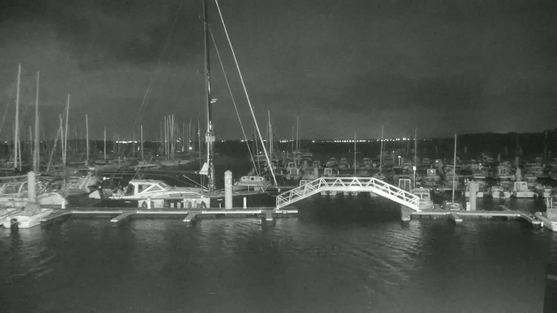 A nighttime, low-light view of a marina filled with numerous boats and sailboats, with a small pedestrian bridge connecting two docks.