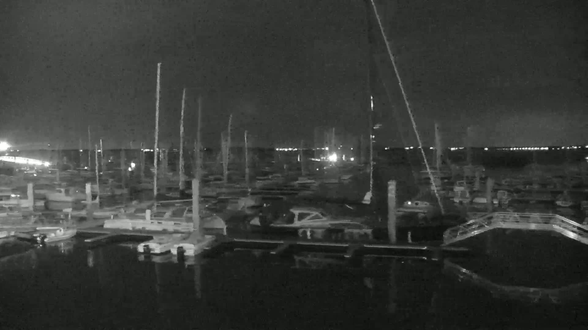 A nighttime, low-light image shows numerous boats docked at a marina, with faint lights visible in the distance across the water.