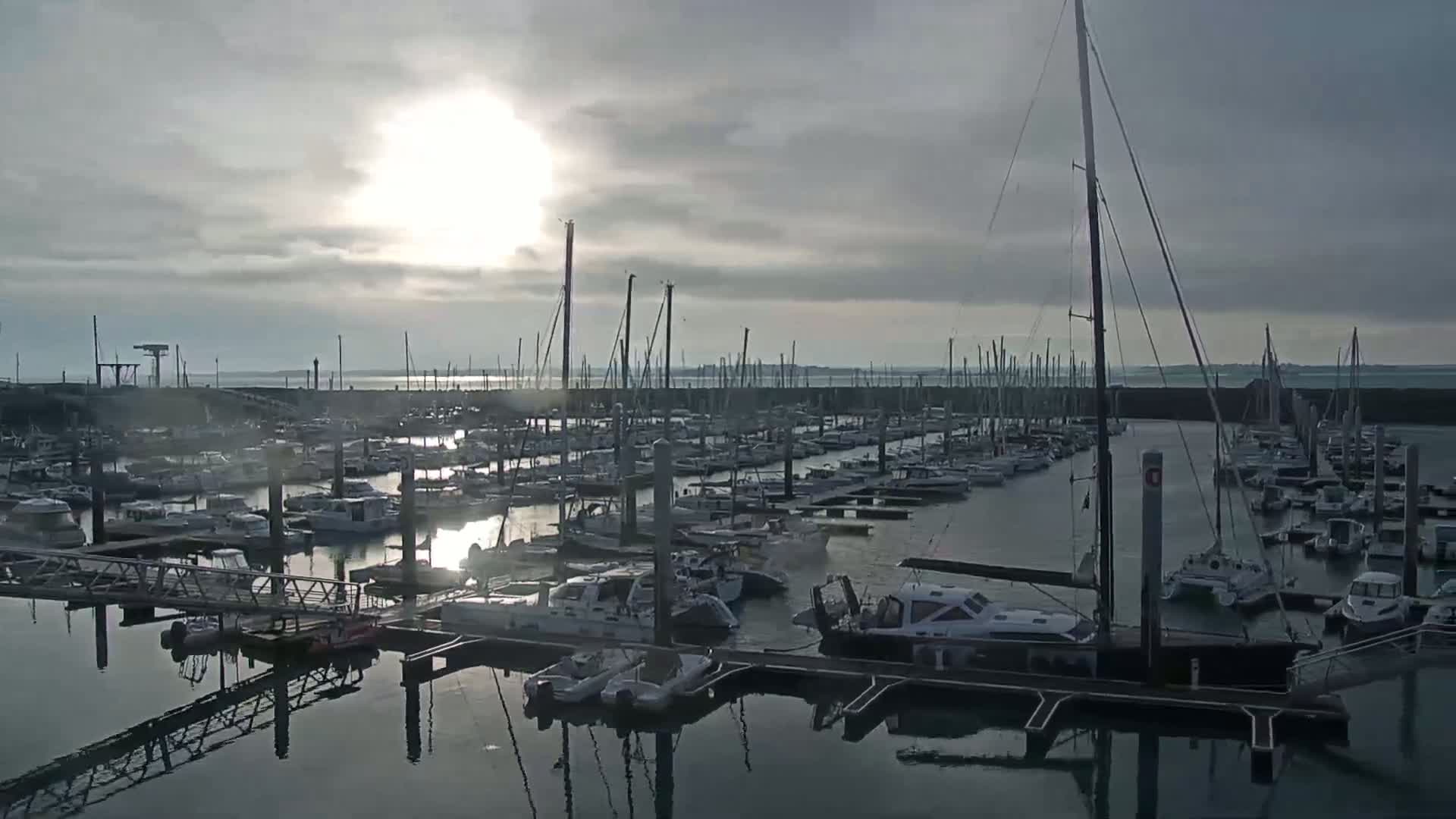 A marina filled with numerous sailboats and motorboats is shown under a cloudy sky with the sun shining through the clouds.