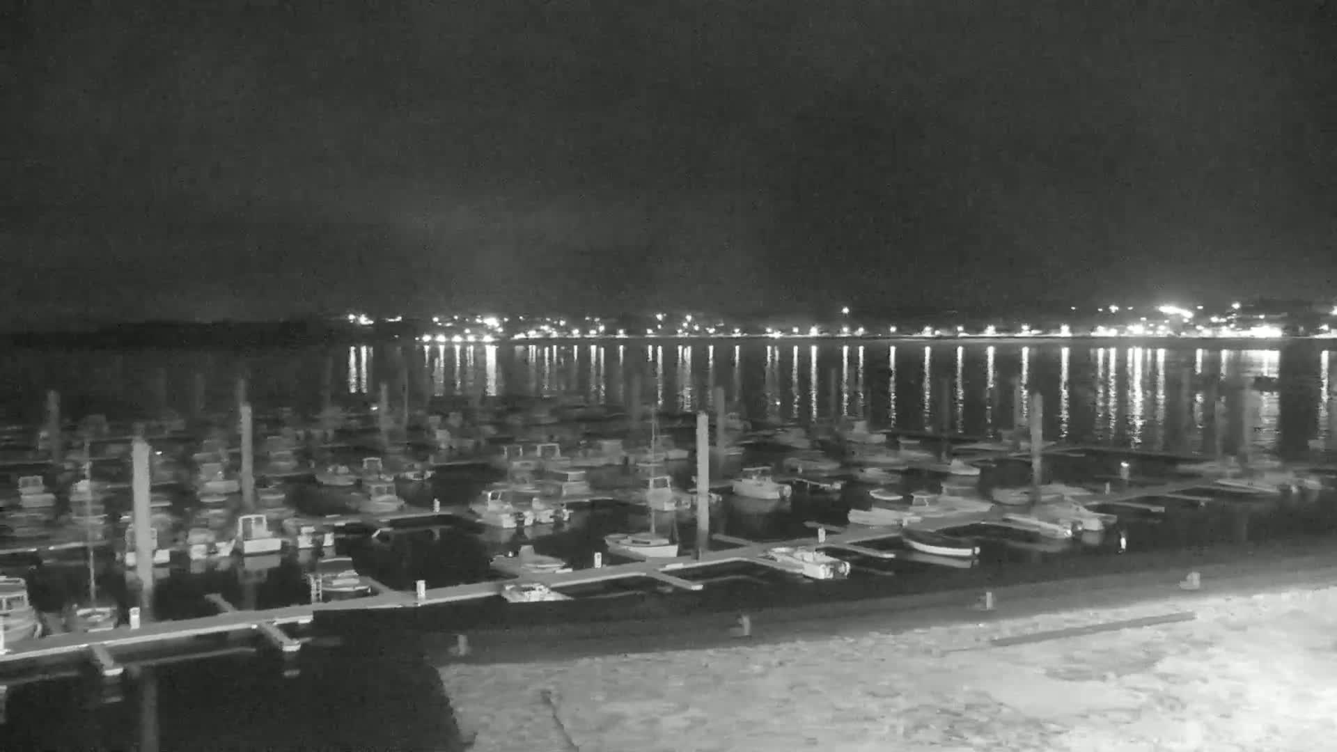 A marina at night with many boats docked, calm water reflecting lights from a distant town.