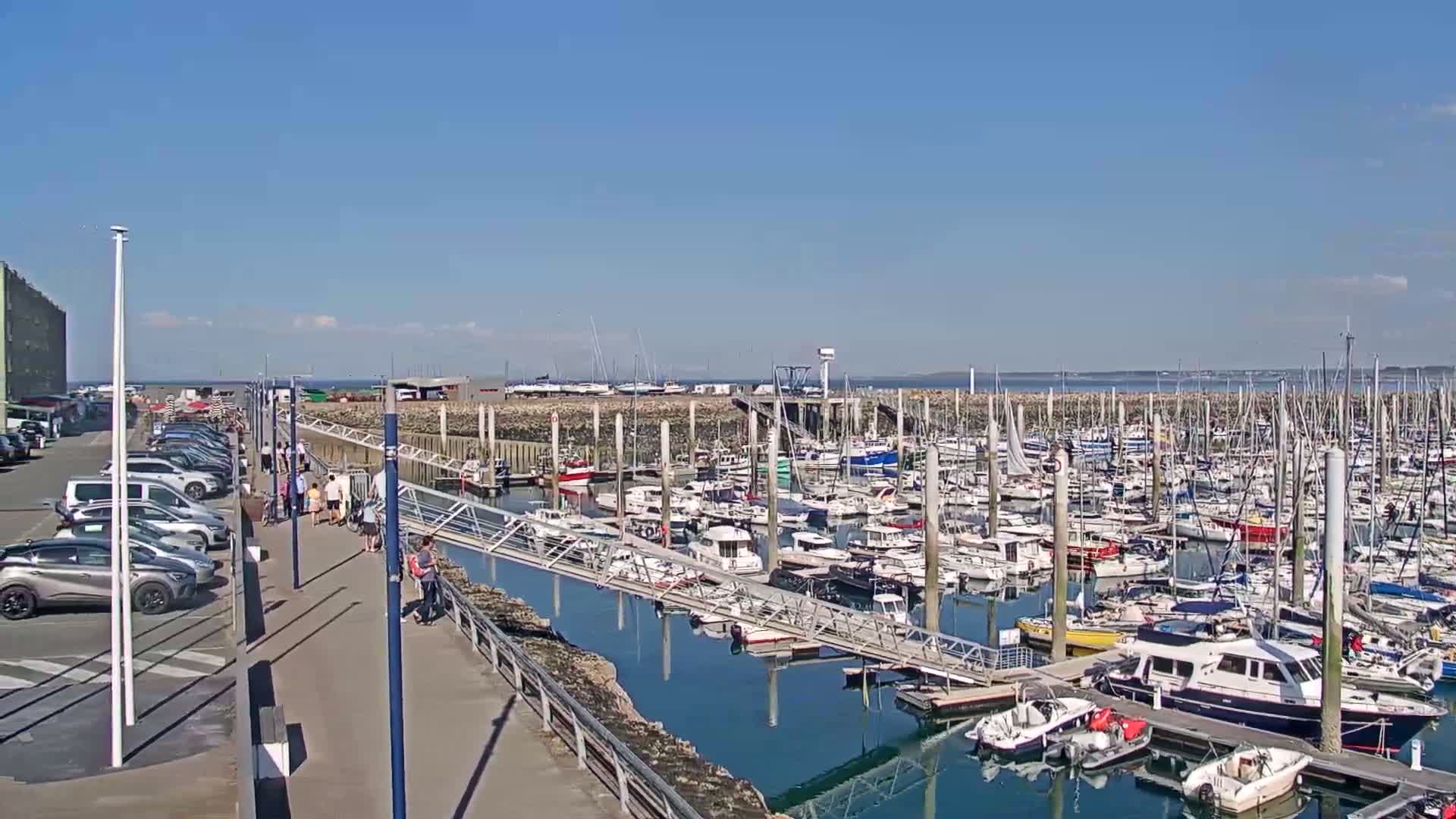 A sunny day shows a marina filled with numerous boats of varying sizes, docked along a walkway with cars parked nearby and people strolling.