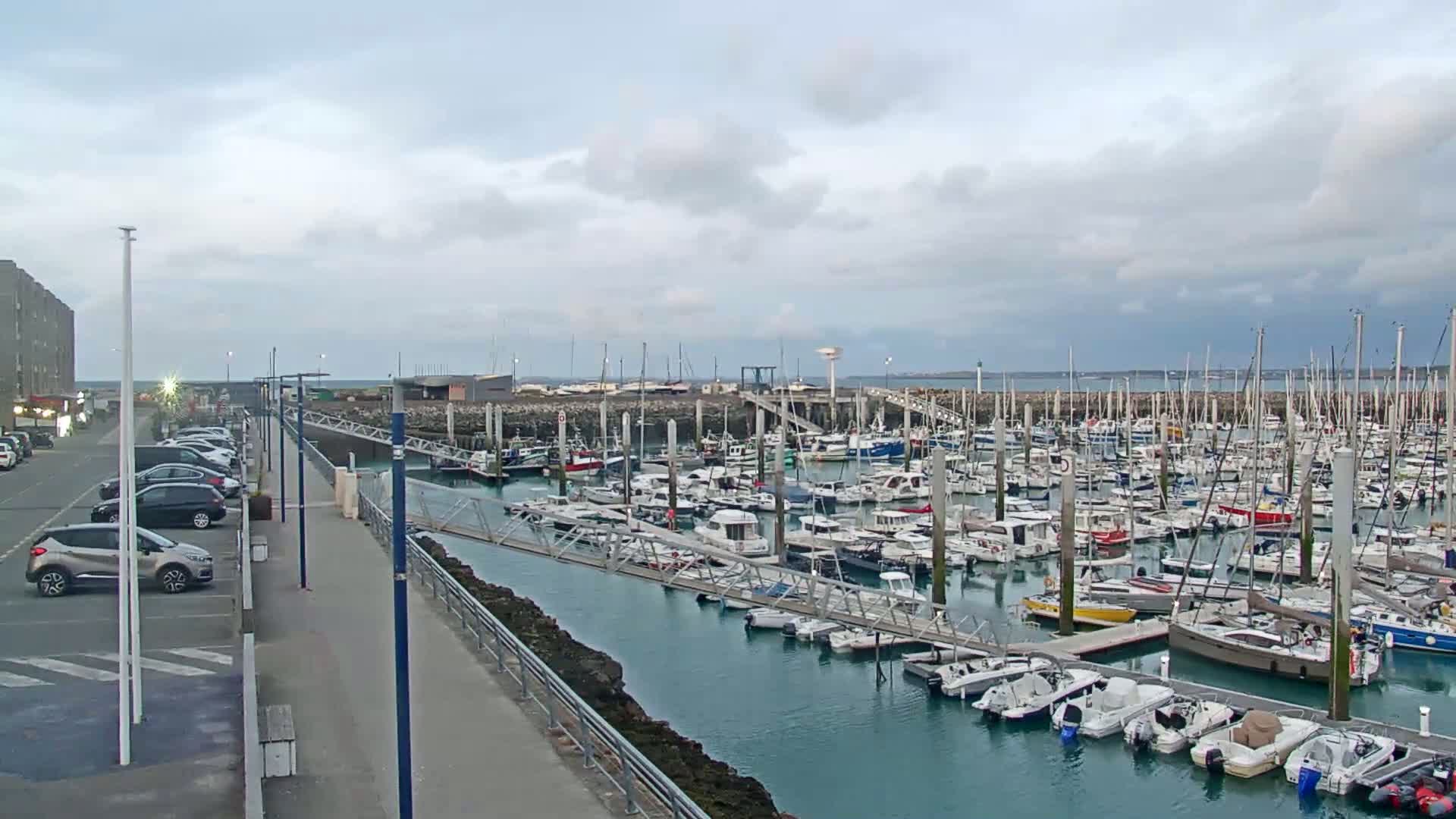 A marina filled with numerous boats is seen under a cloudy sky, next to a parking lot and walkway.