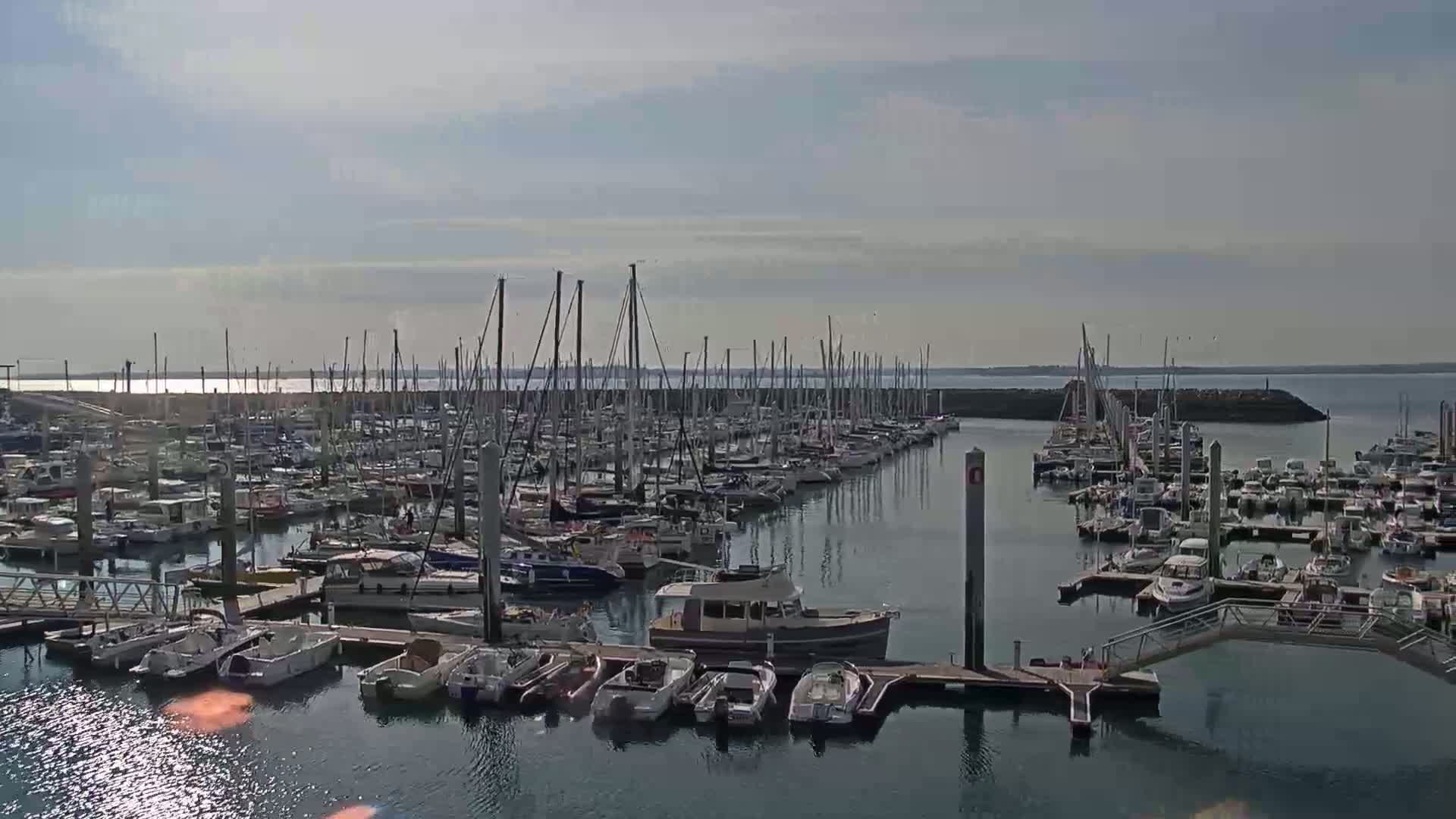 A calm harbor filled with numerous sailboats and motorboats under a mostly sunny sky.