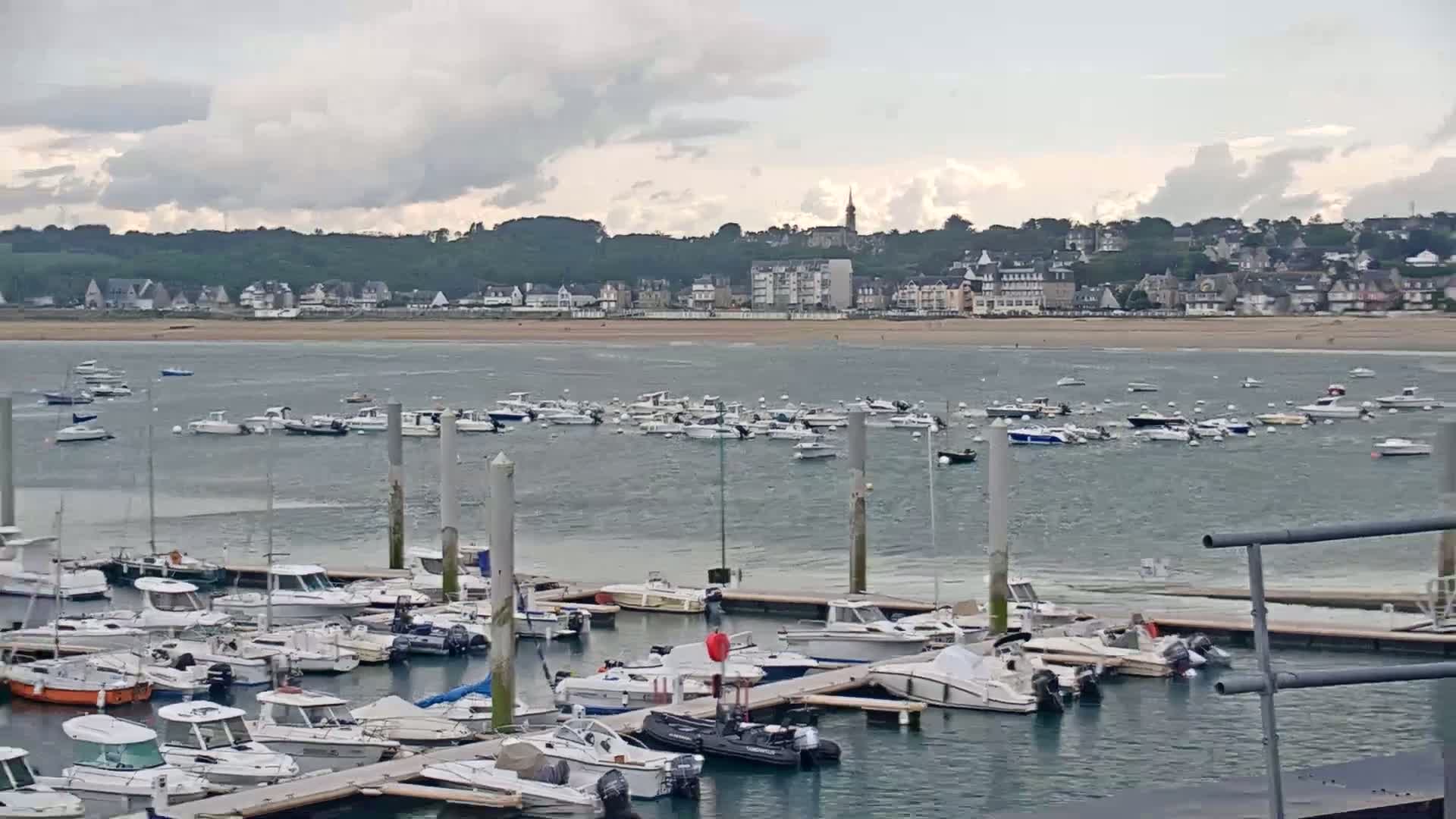 Many boats are moored in a marina, with numerous other boats anchored in the bay beyond, under a cloudy sky.