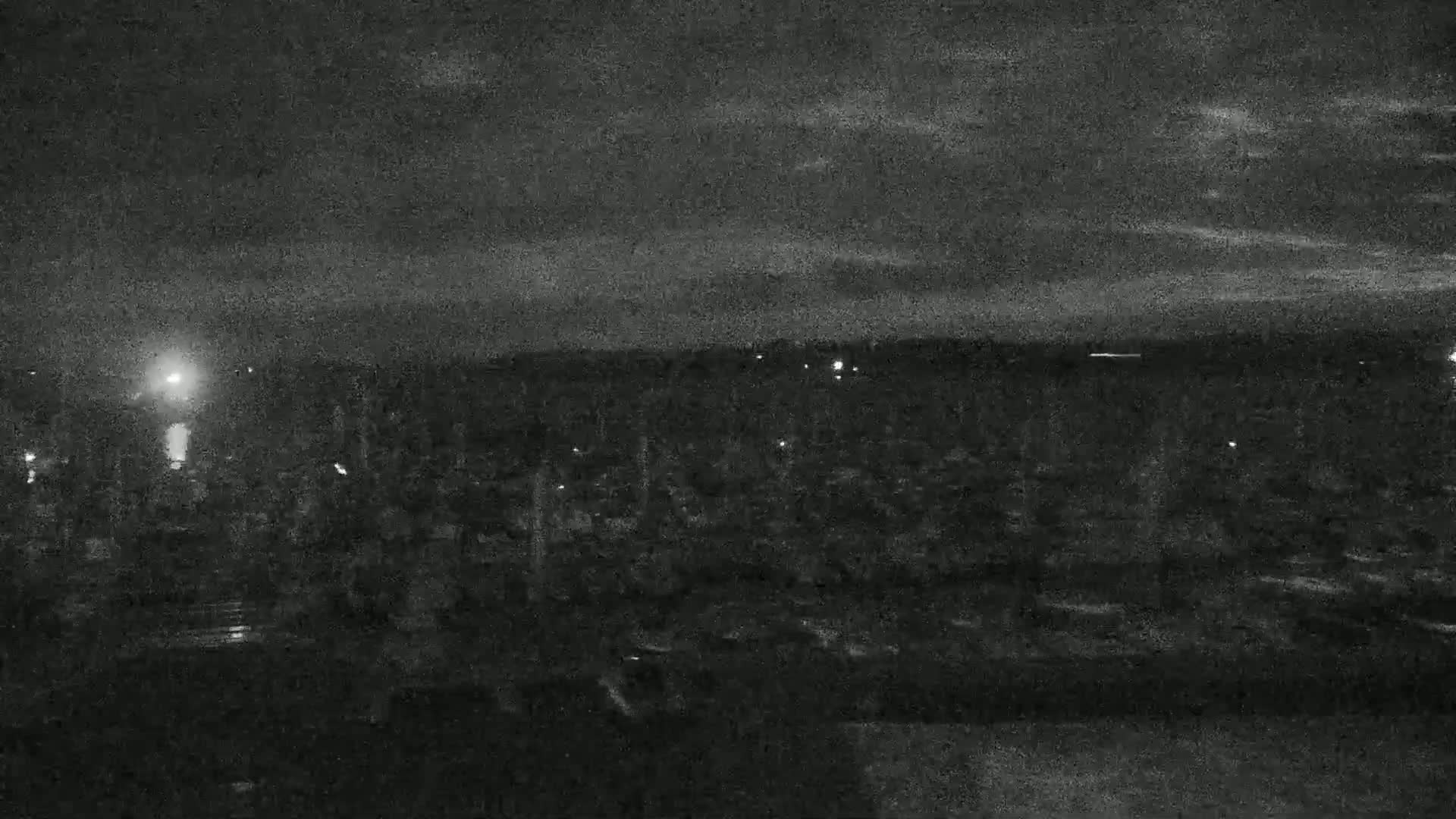A nighttime, low-light view shows a dark landscape with sparsely scattered lights under a cloudy sky.