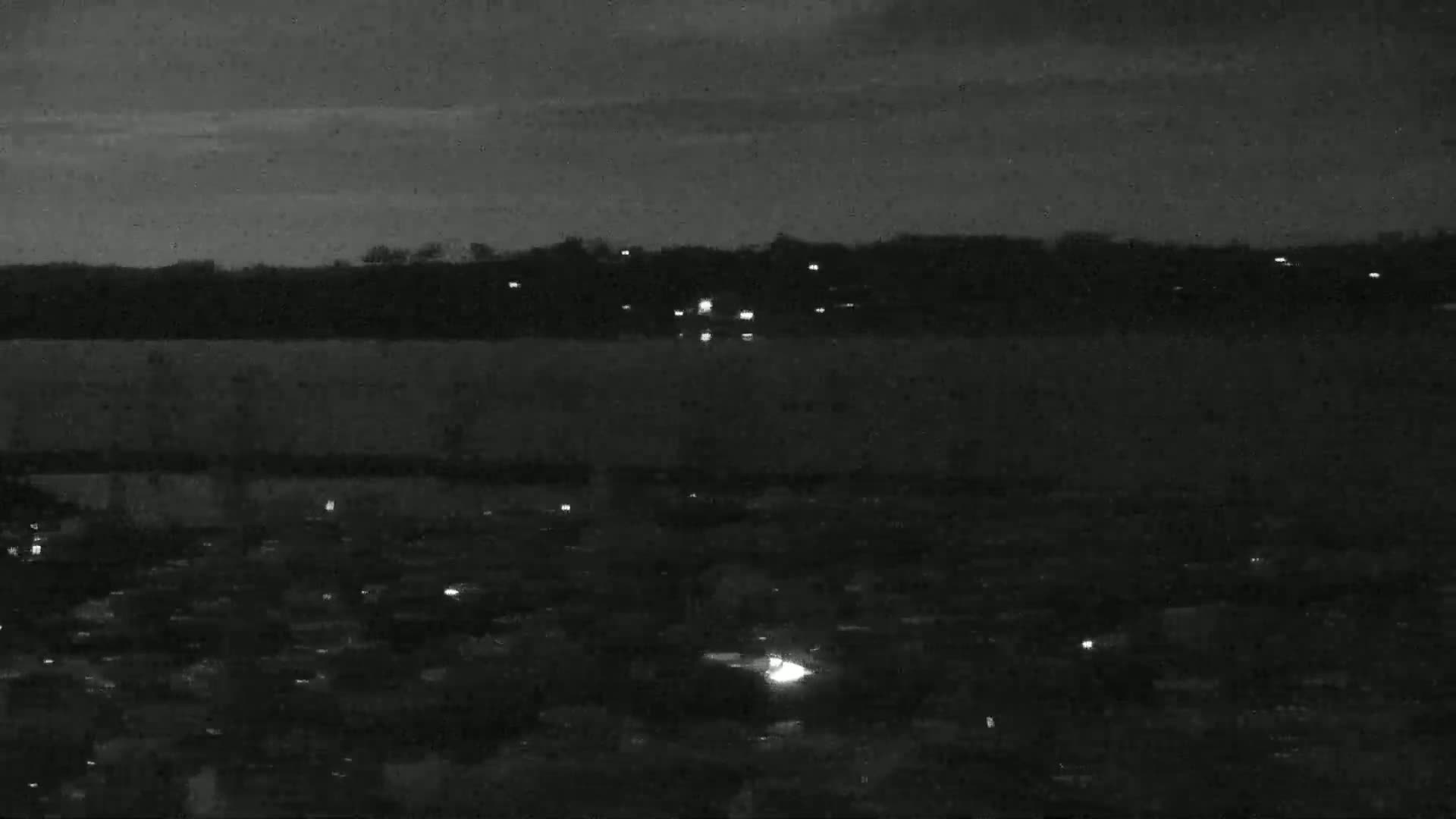 A nighttime view shows a body of water with sparse lights on the far shore and a residential area with scattered lights in the foreground under a dark, cloudy sky.