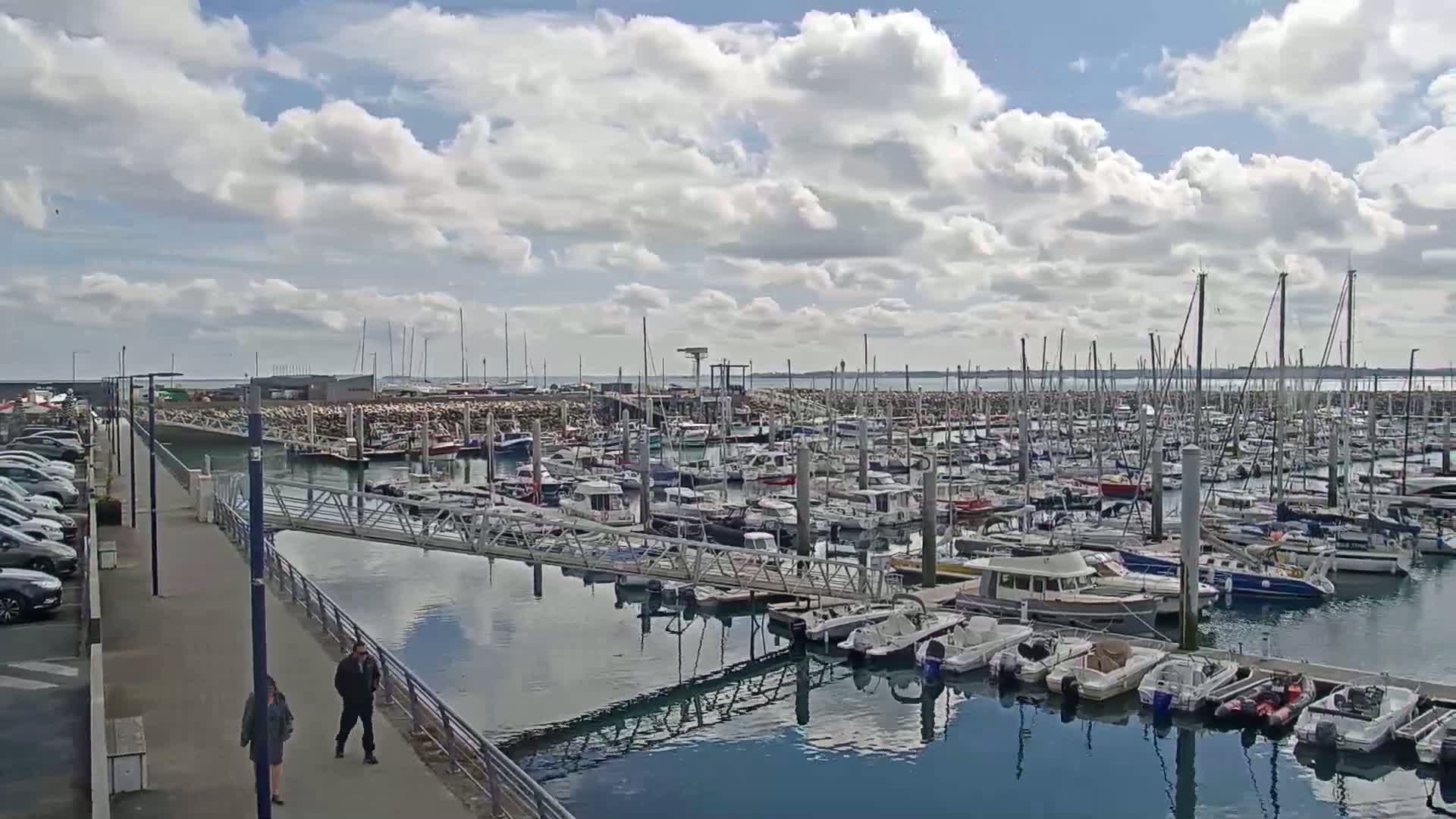 A marina filled with numerous boats and yachts under a partly cloudy sky, with two people walking along a paved walkway beside the water.
