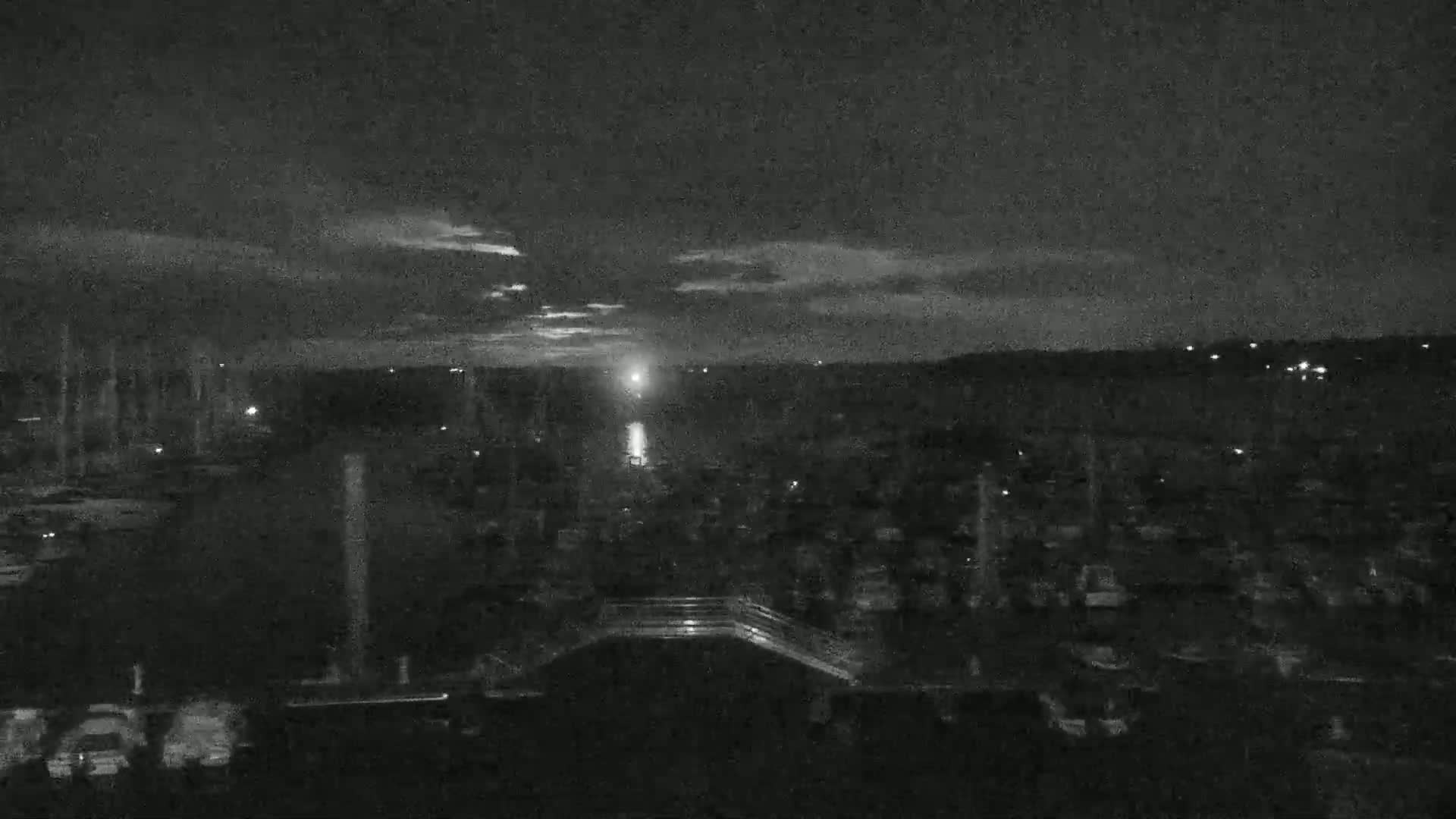 A nighttime, low-light view of a marina with many boats, a bridge, and a bright light in the distance under a partly cloudy sky.