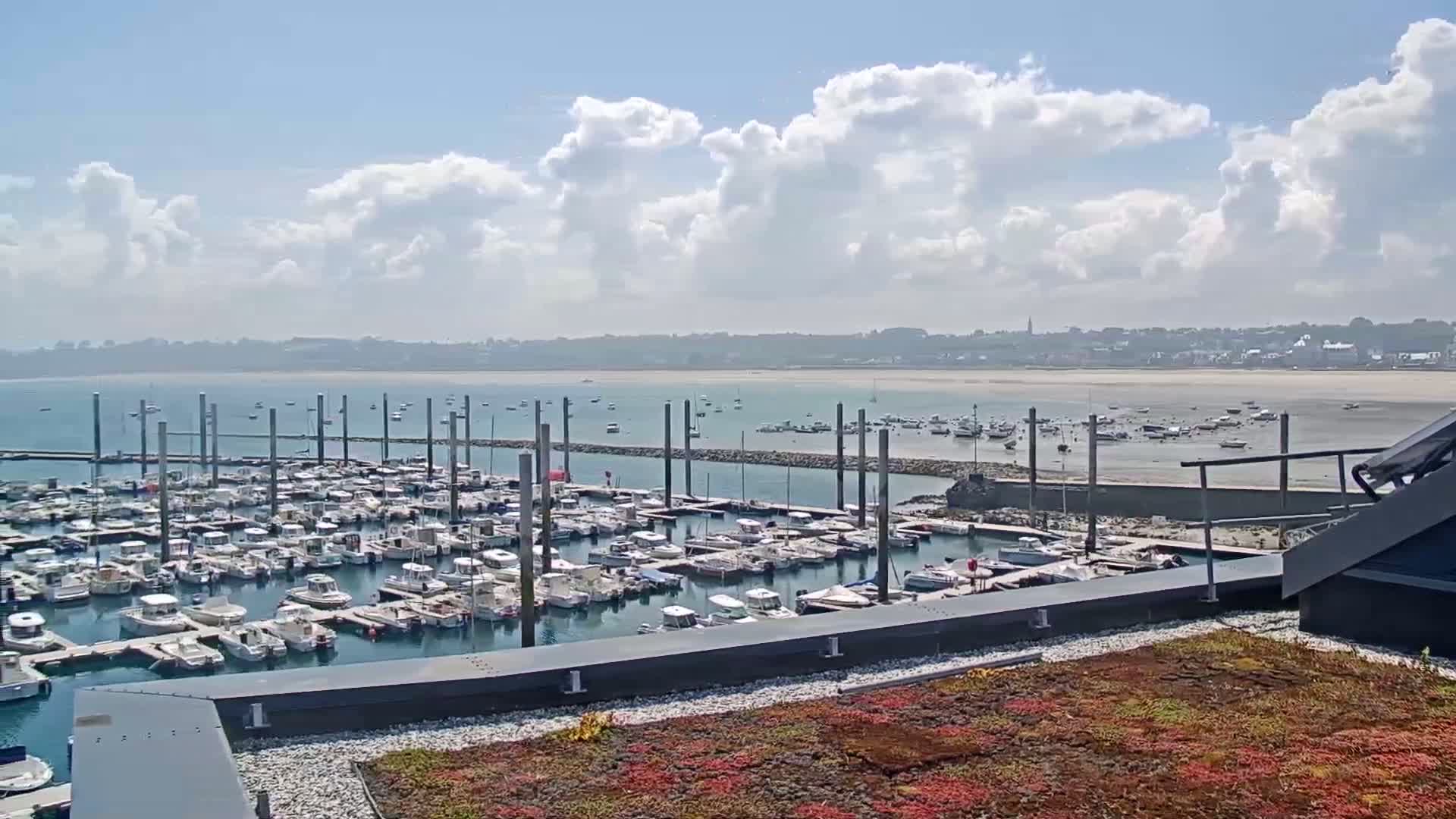 A marina filled with numerous boats is situated near a sandy beach and town under a partly cloudy sky.