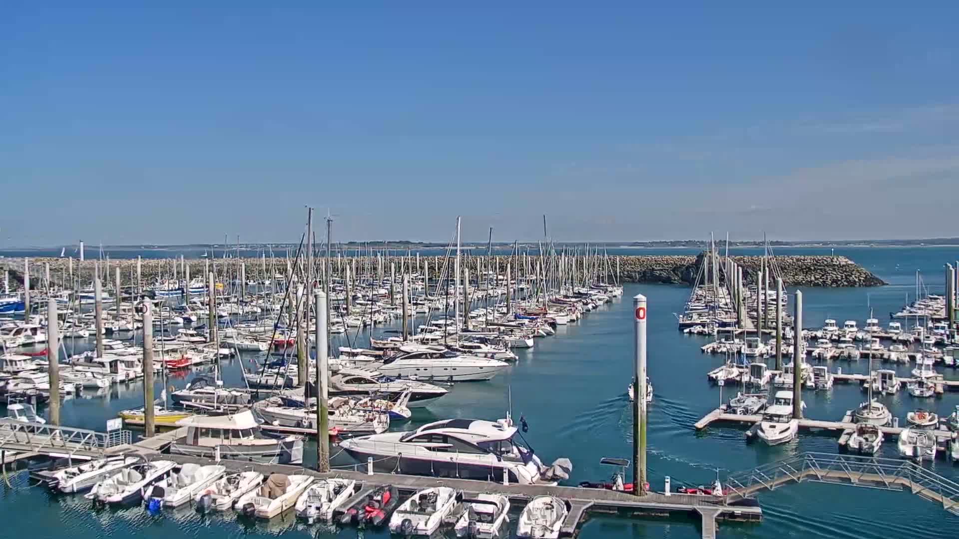 A large marina filled with numerous sailboats and motorboats is shown on a sunny day with calm water.