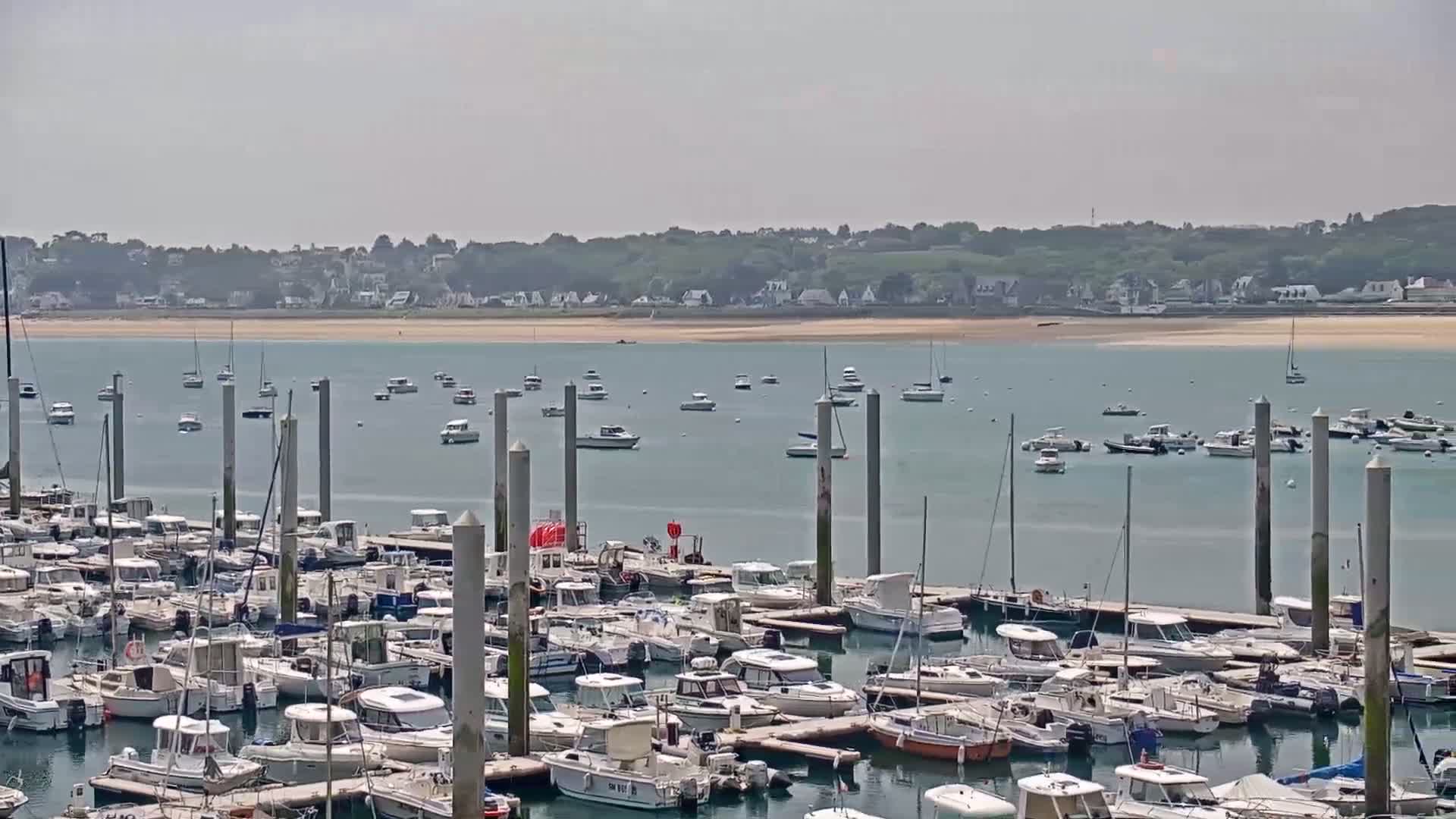 Many boats are docked in a marina, with additional boats anchored in the calm water beyond, under a hazy sky.